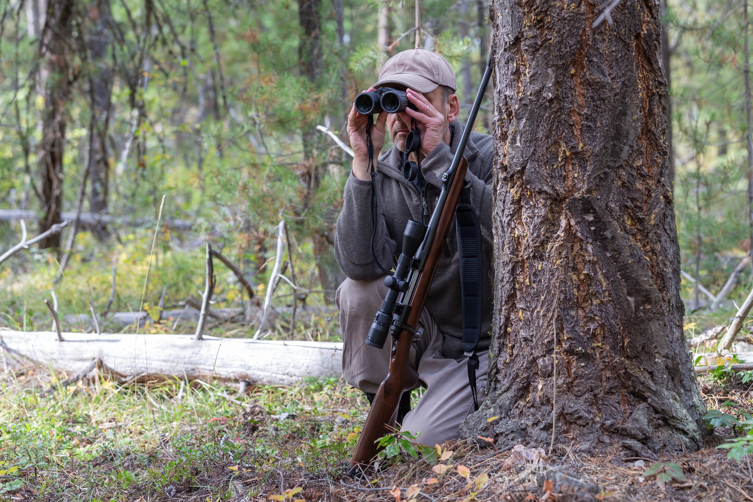 Hunter hiding behind a tree, watching the surroundings with binoculars. A rifle with the scope is leaning against the tree in front of him.