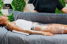 Person undergoing a body wrap treatment on a spa bed, covered in a white substance, attended by a professional.