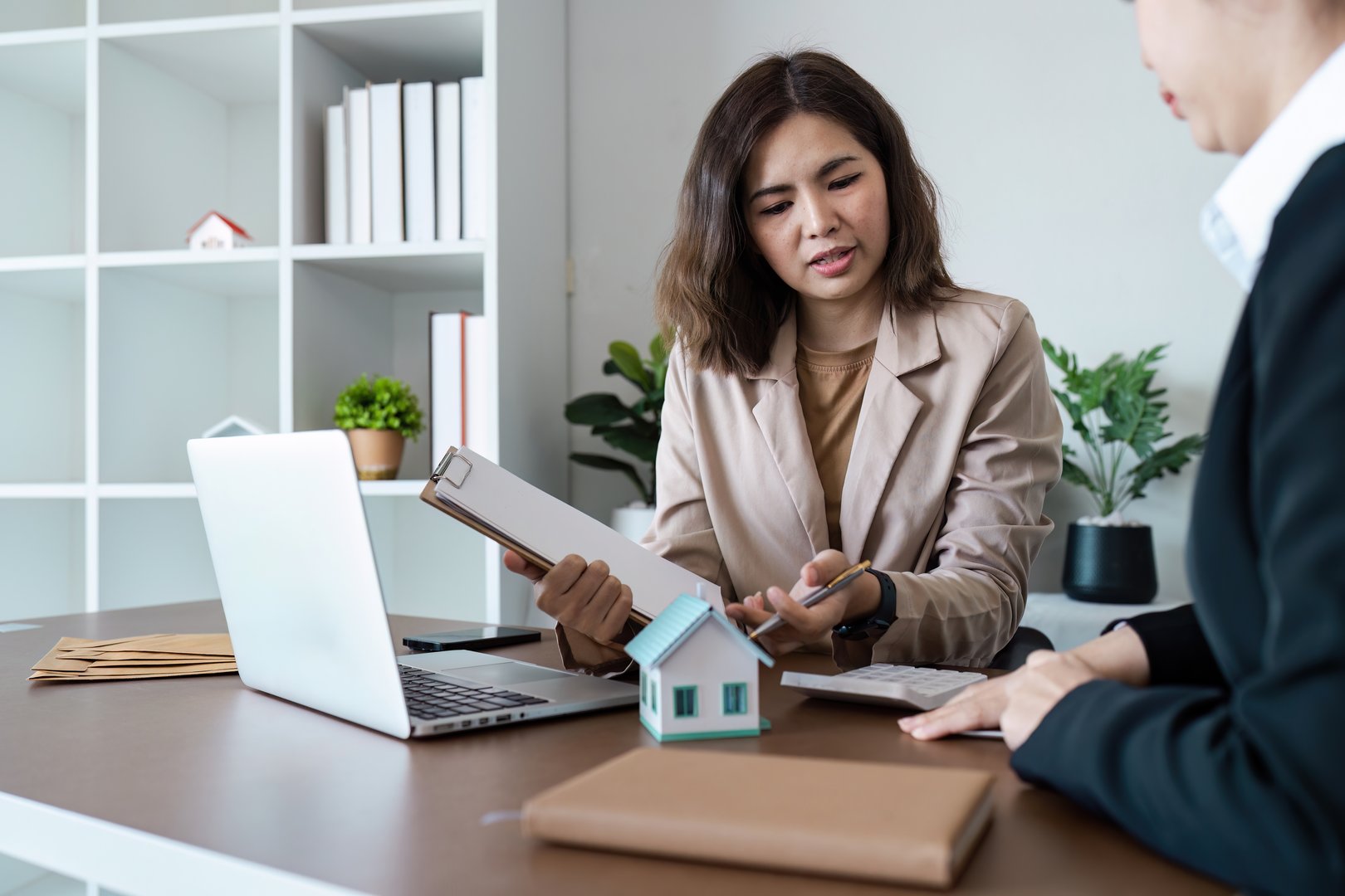 Real estate agent explaining property details to a client in a modern office, showcasing a model house and documents.