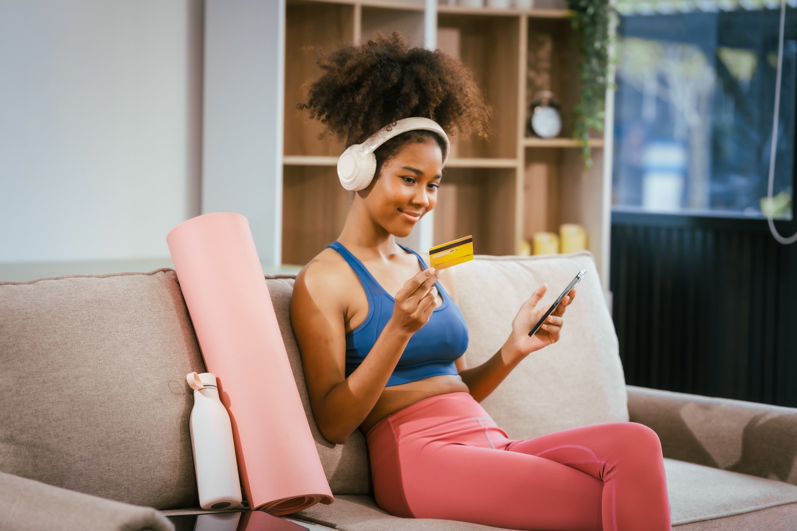 A young African American woman in workout clothes sits on the couch at home after a workout, smiling while looking at her computer, online shopping with a credit card in hand