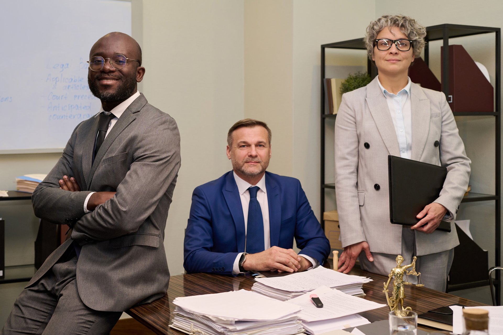 Young Black man standing with arms crossed next to middle aged Caucasian woman holding folder and middle aged Caucasian man sitting at desk, with documents in office setting