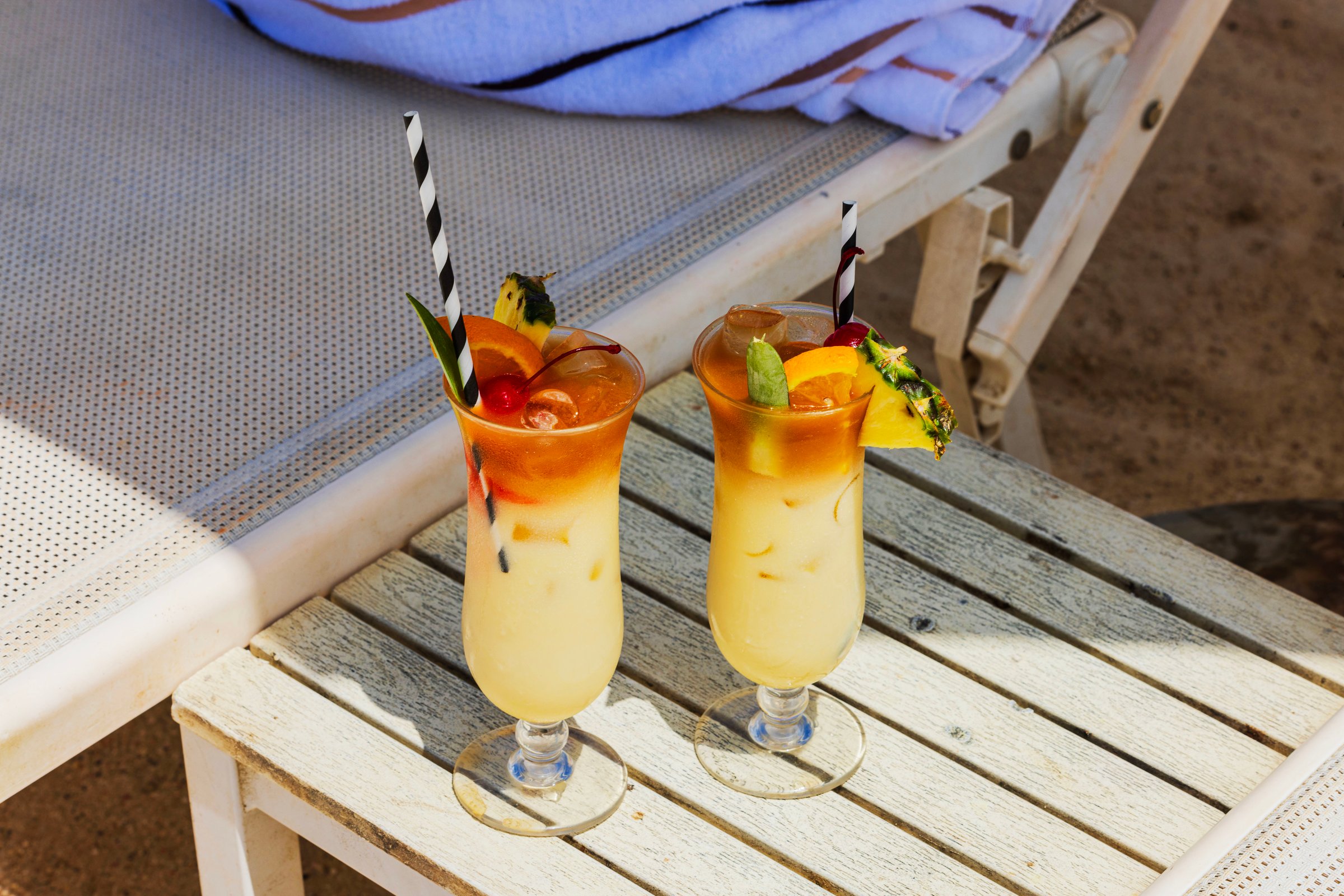 Two tropical Mai Tai cocktails with pineapple and cherry garnish served on by sun lounger at beach. Curacao.