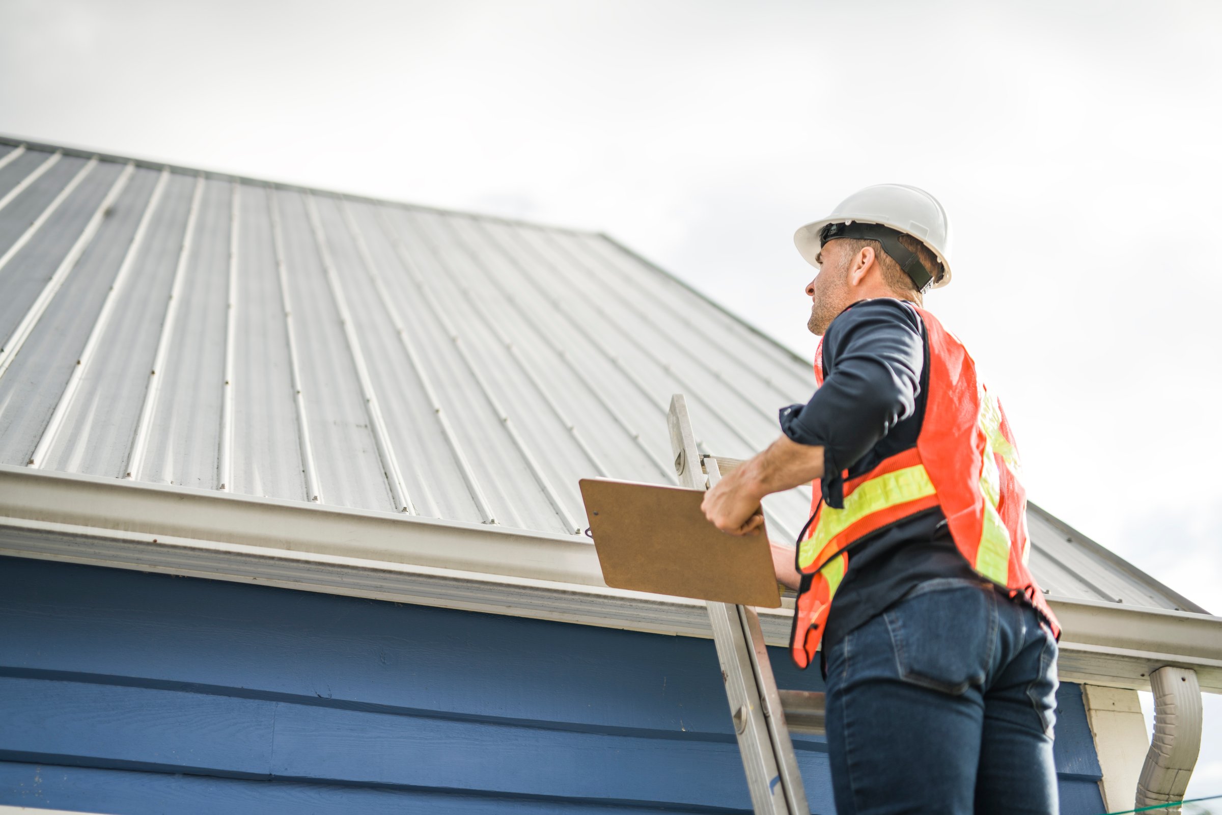 A man with hard hat standing on steps inspecting house roof