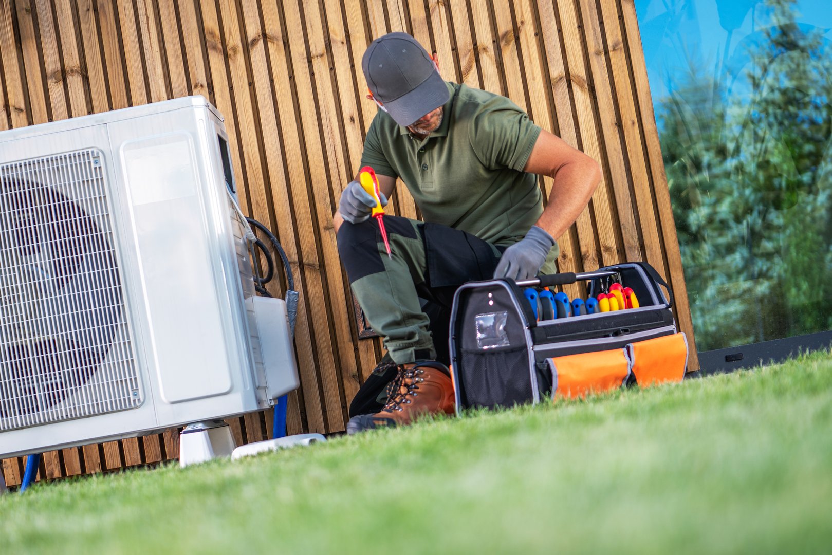 A technician repairs an air conditioning unit outside, meticulously organizing tools in a bag while seated on the grass.