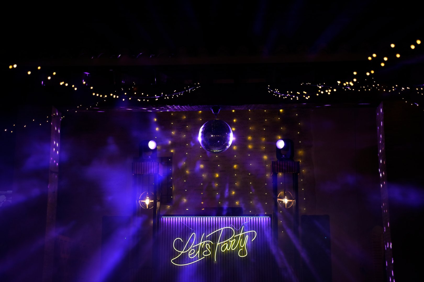 A disco ball hangs from the ceiling of a club. The club is called Let's Party. The lights are purple and the club is lit up