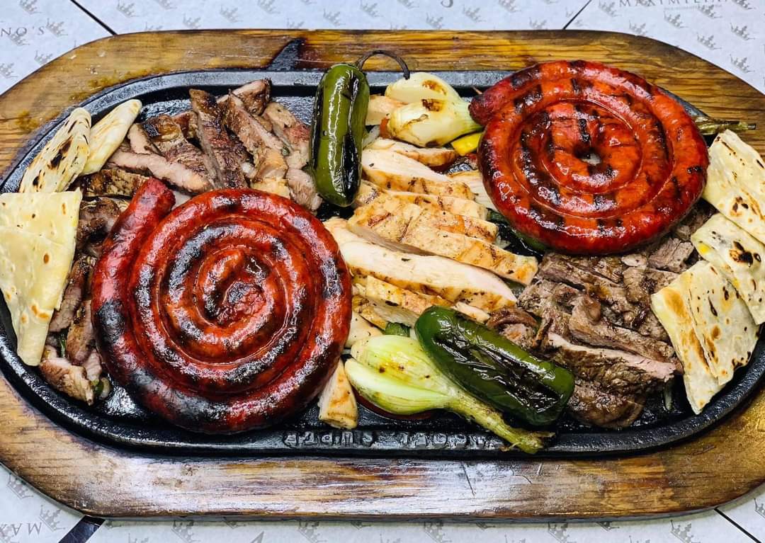 Grilled meat platter with sausages, sliced beef, chicken, onions, jalapeños, and tortillas on a sizzling plate.