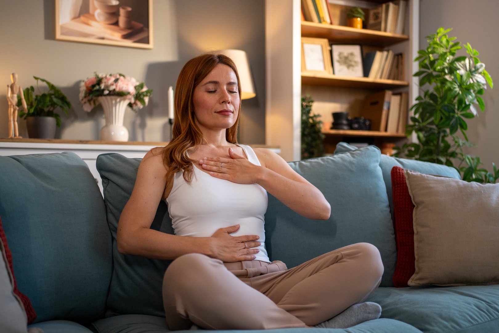 Young adult woman sitting in lotus position on a comfortable sofa, practicing diaphragmatic breathing with closed eyes, promoting relaxation and mindfulness in her living room