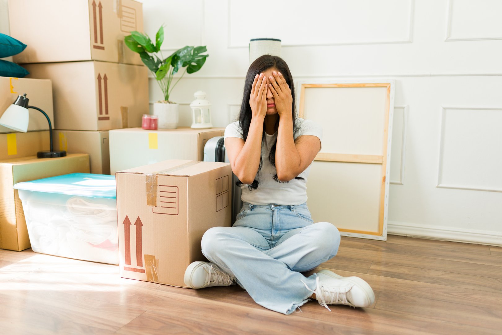 Exhausted woman covering her face with her hands while sitting on the floor surrounded by cardboard boxes and feeling unable to unpack in her new apartment