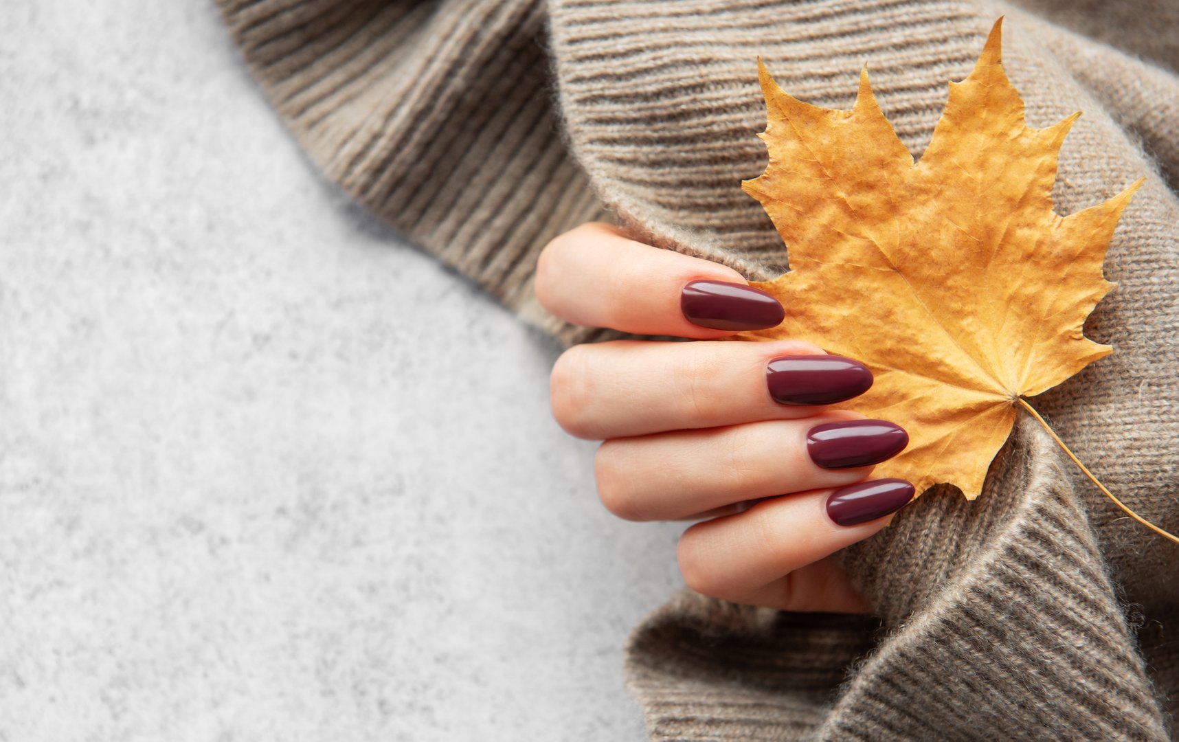 A hand with elegant dark nails gently holds a bright orange maple leaf against a soft beige sweater. The cozy atmosphere captures the essence of autumn.