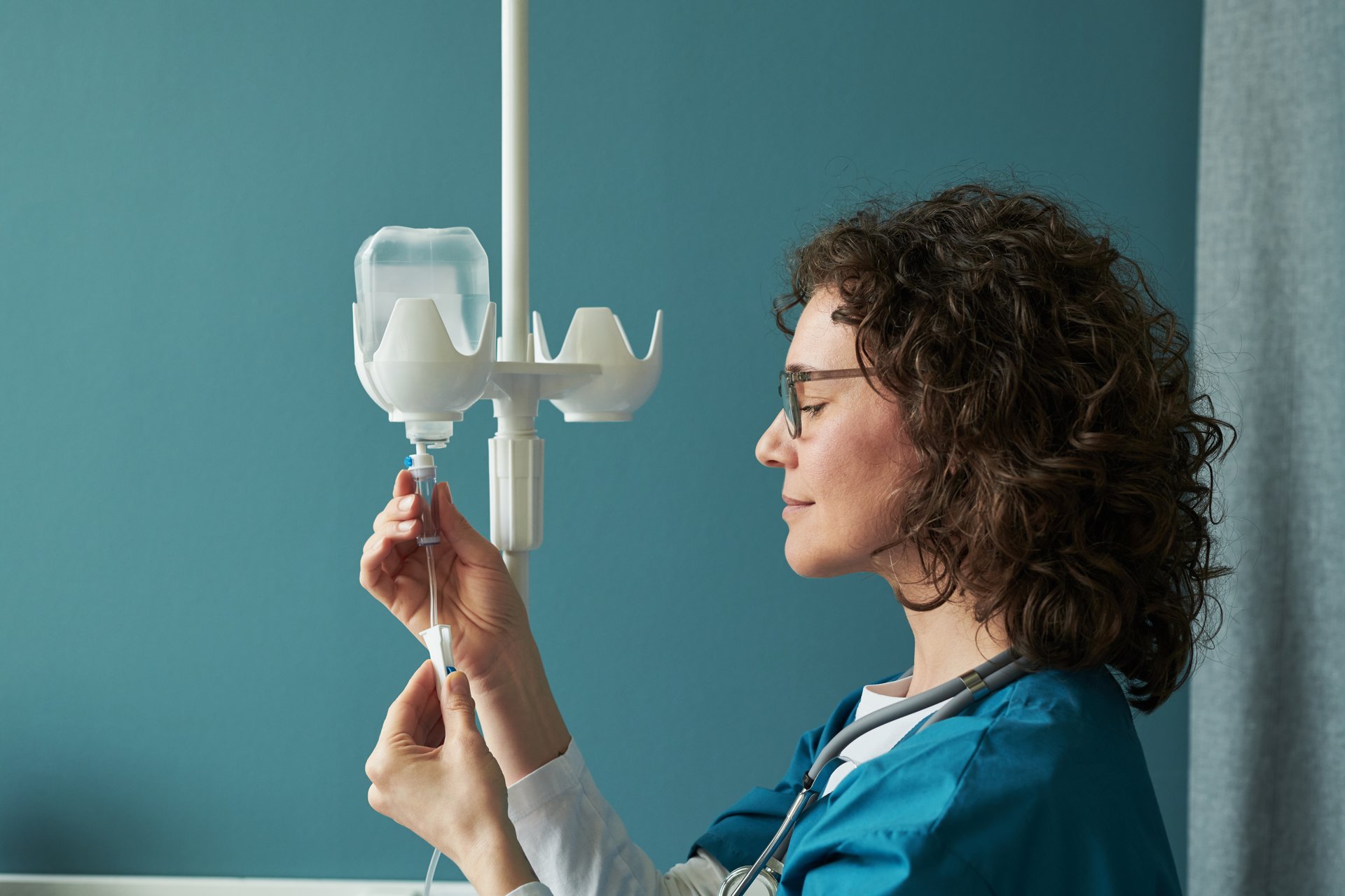 Middle aged Caucasian woman nurse preparing intravenous drip, adjusting IV line with focused expression, wearing medical scrubs and stethoscope in clinical setting