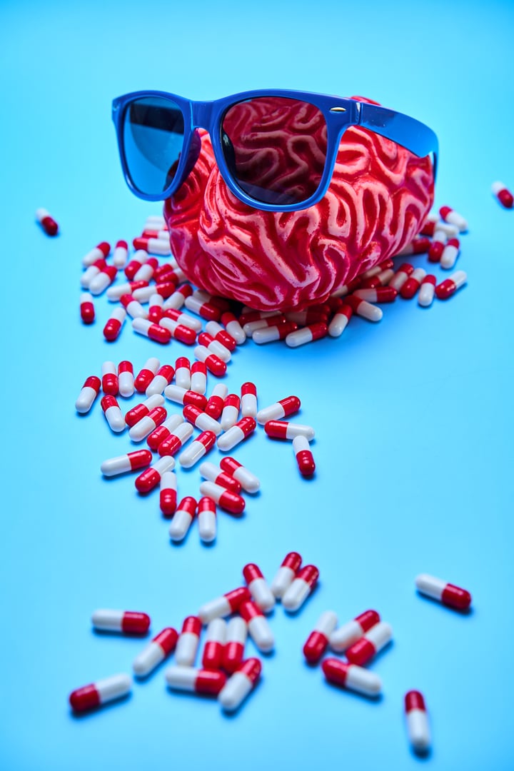 Red brain with sunglasses on top of a pile of red and white capsules on a blue surface.