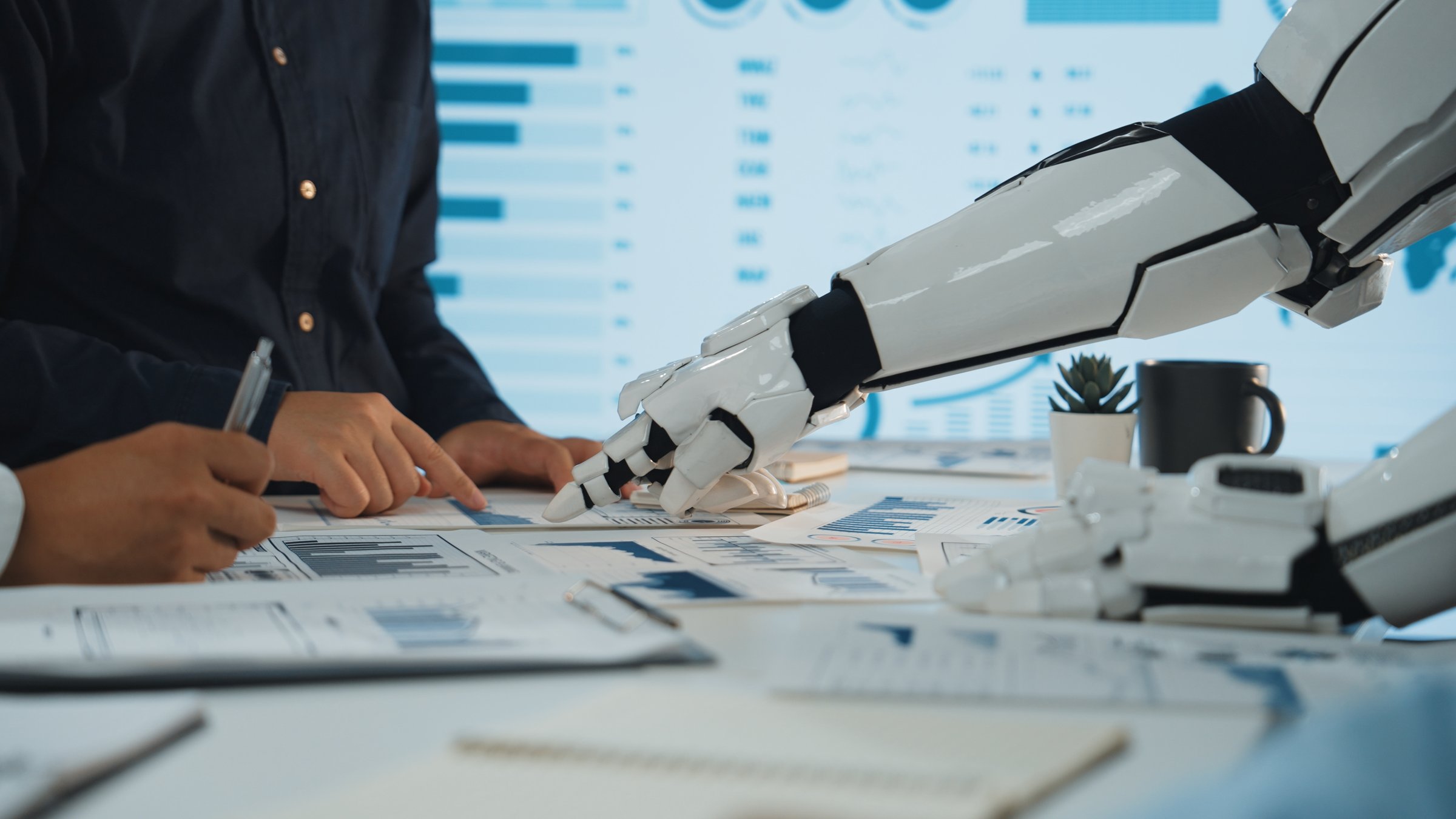 A modern office scene showcasing collaboration between humans and robots. A robotic arm assists with data analysis, highlighting technology integration in business environments.Noogenesis