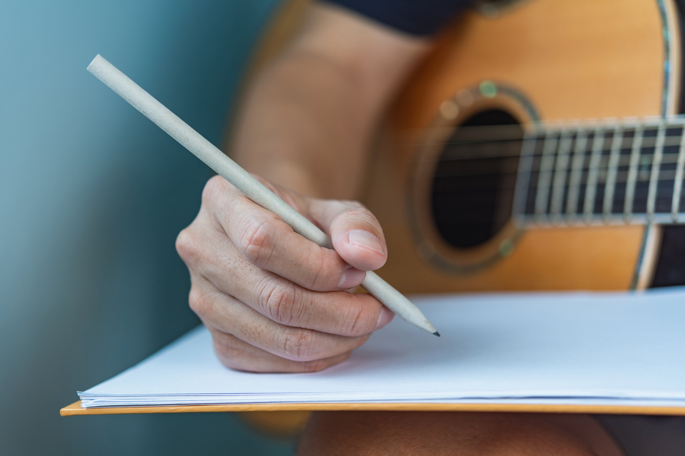 Close-up of a songwriter's hand writing lyrics with a guitar in the background.