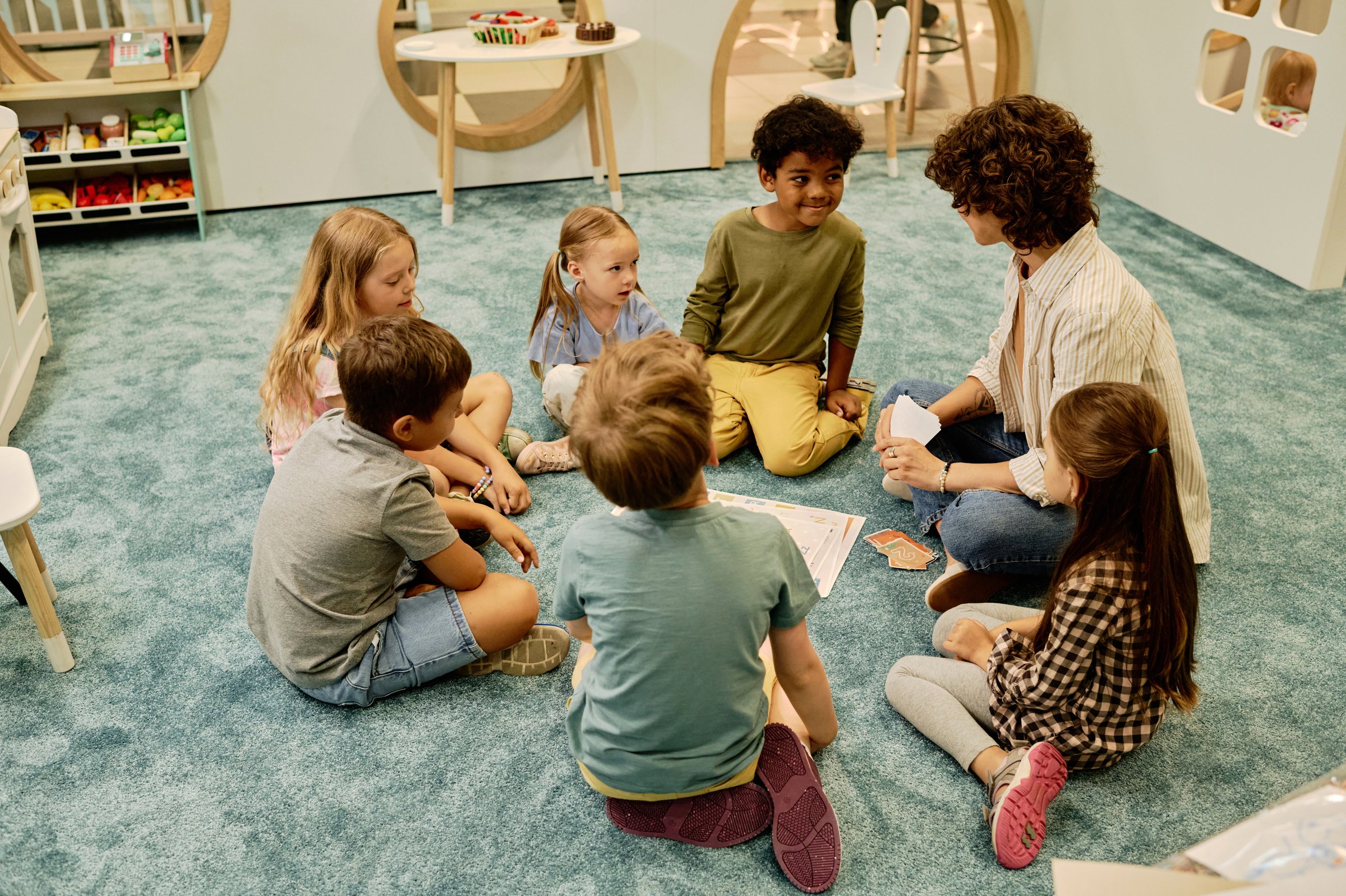 Group of cute intercultural kids sitting in circle on the floor and looking at young female teacher during discussion of topic