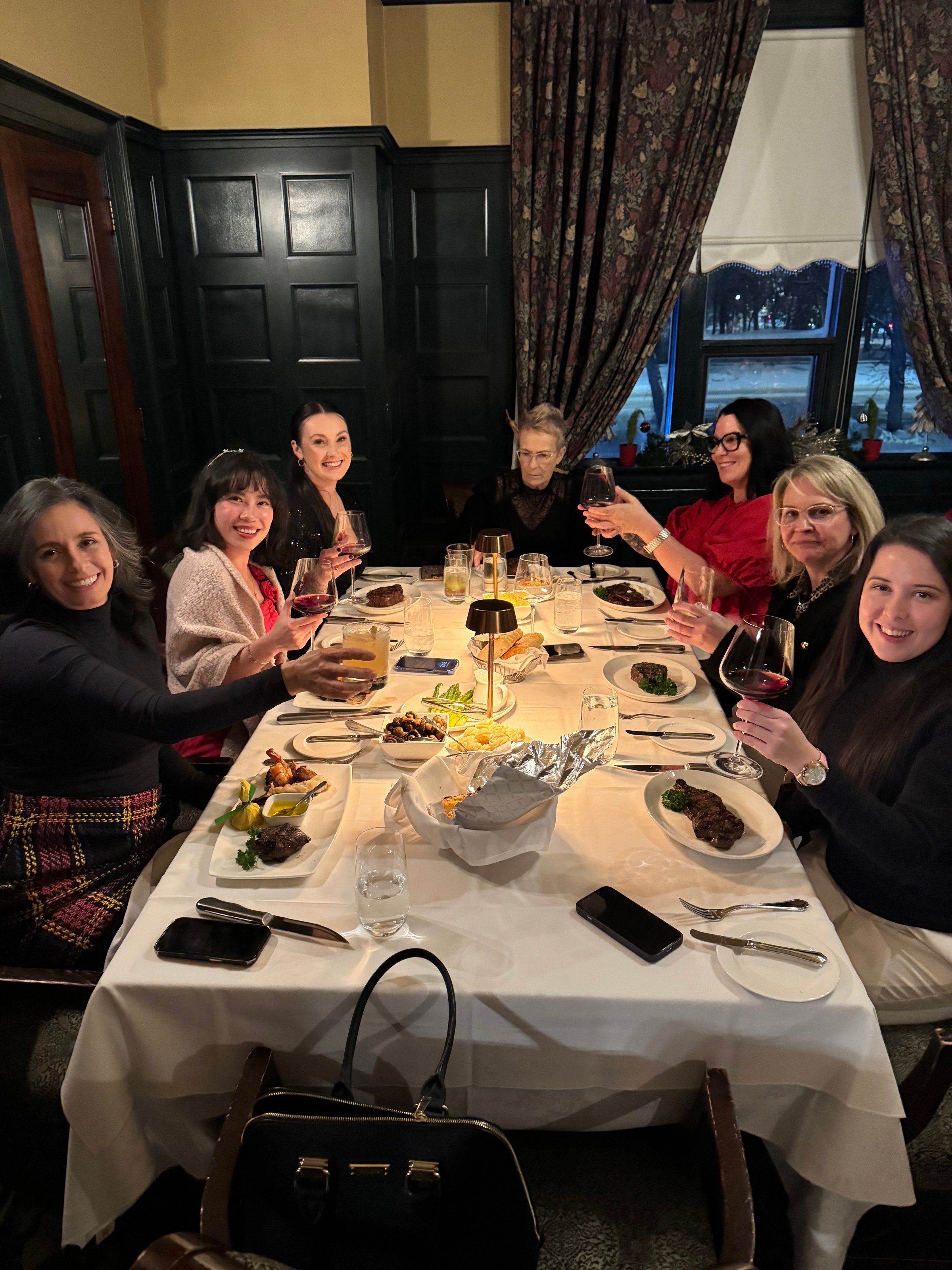 A group of seven women sitting around a table in a restaurant, toasting with wine glasses and enjoying a meal.