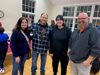 Four people smiling and posing indoors; two men and two women wearing casual clothing in a room with large windows.
