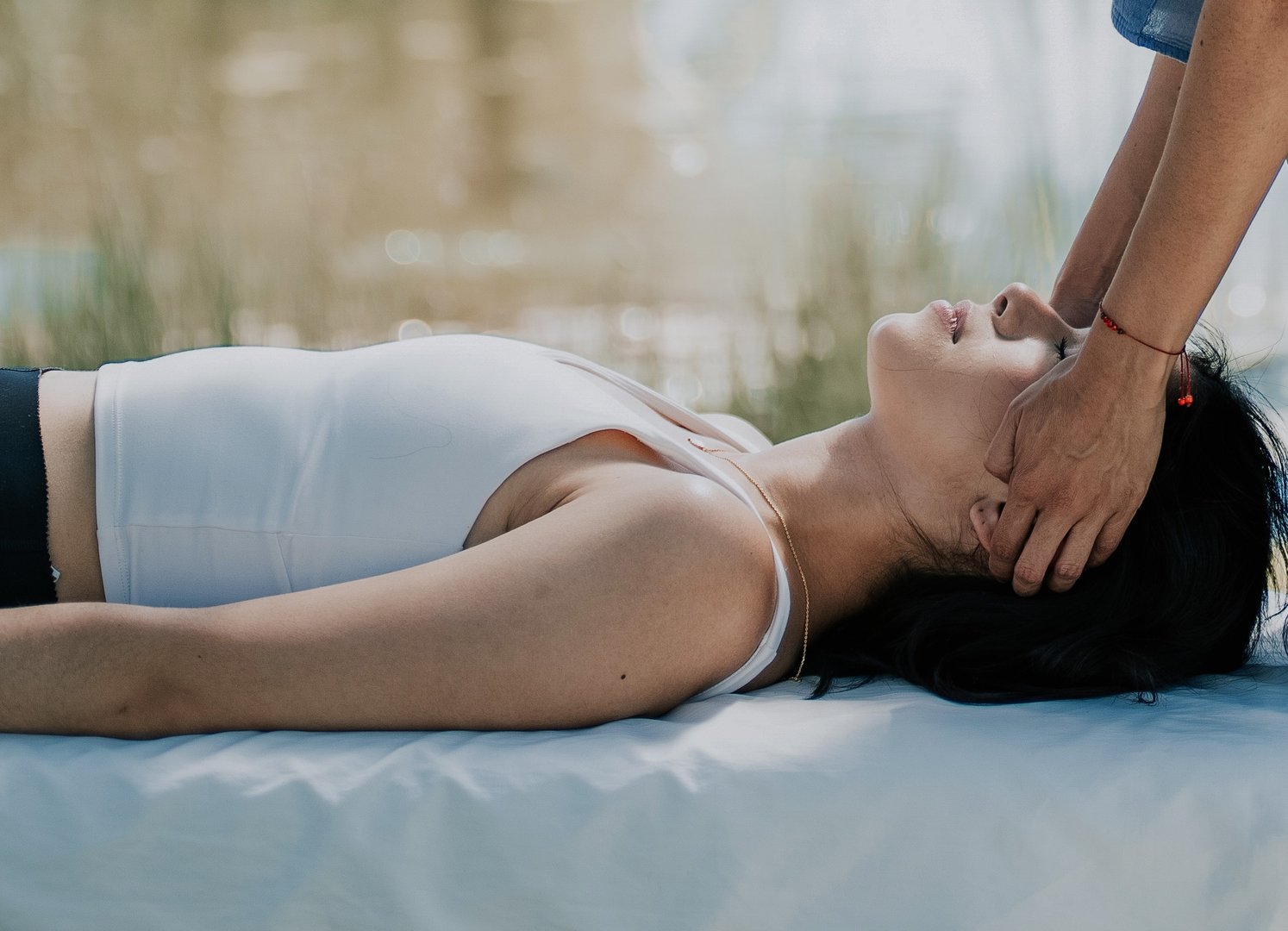 A person lies on a massage table outdoors while another person gently holds their head, providing relaxation therapy.