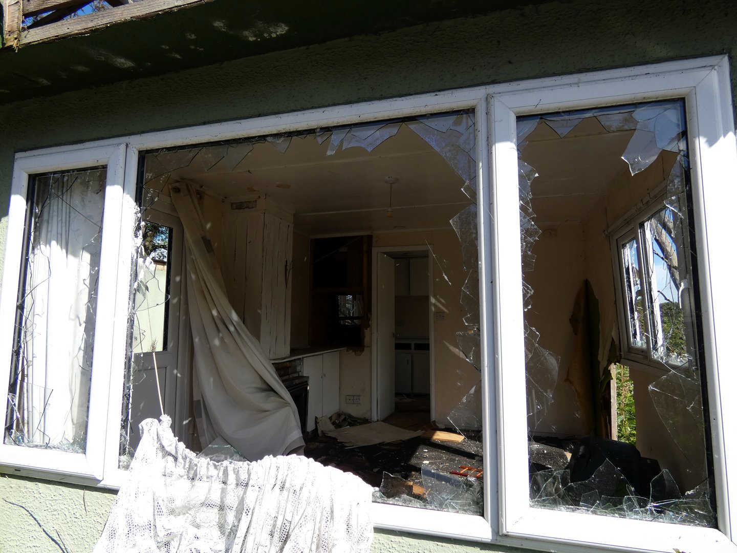 Close-up of a broken window on a pale green mobile home, with cracked glass and peeling paint, suggesting years of neglect