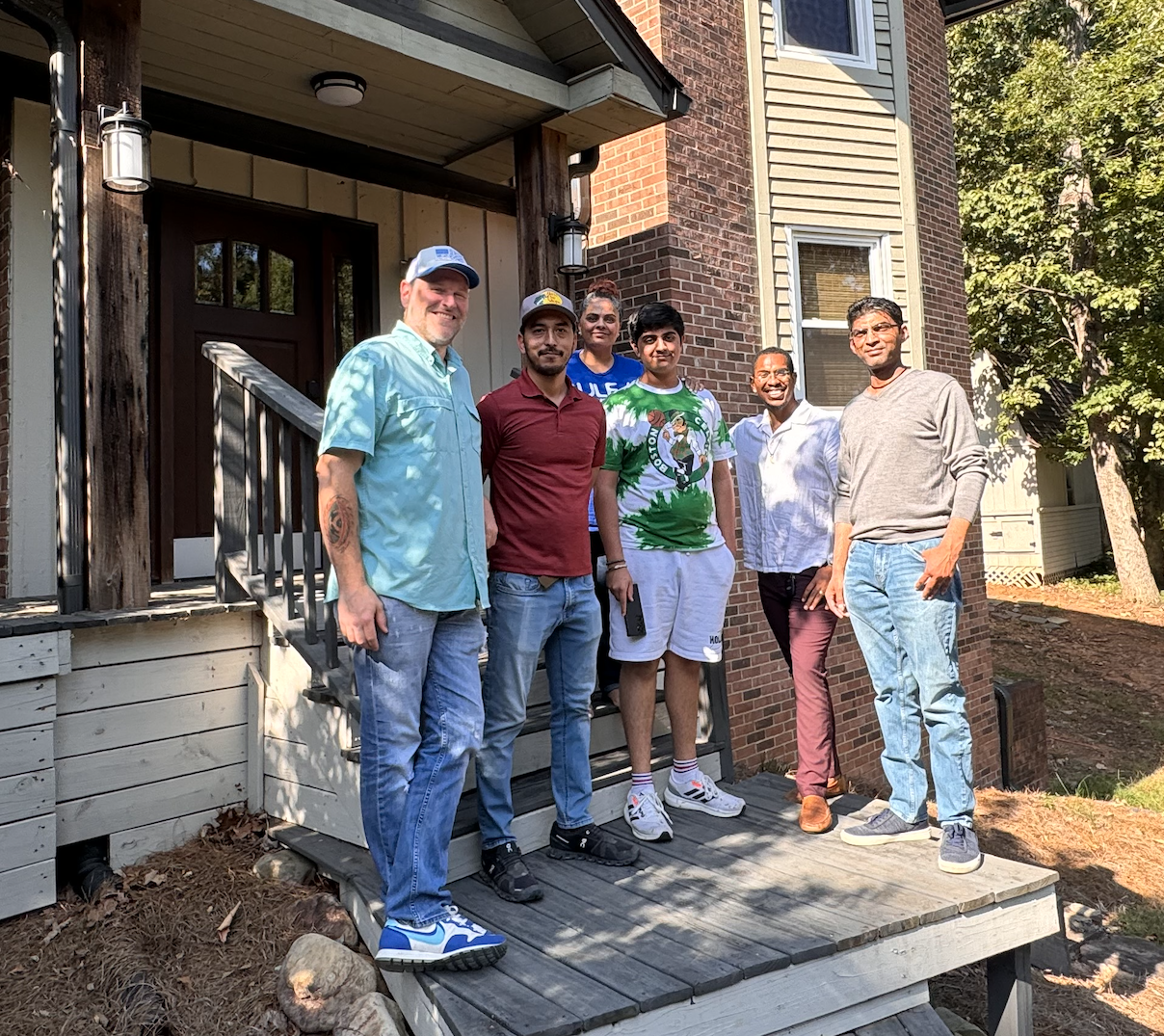 Six men standing on a wooden porch in front of a brick and wood house, wearing casual clothes, and smiling at the camera.