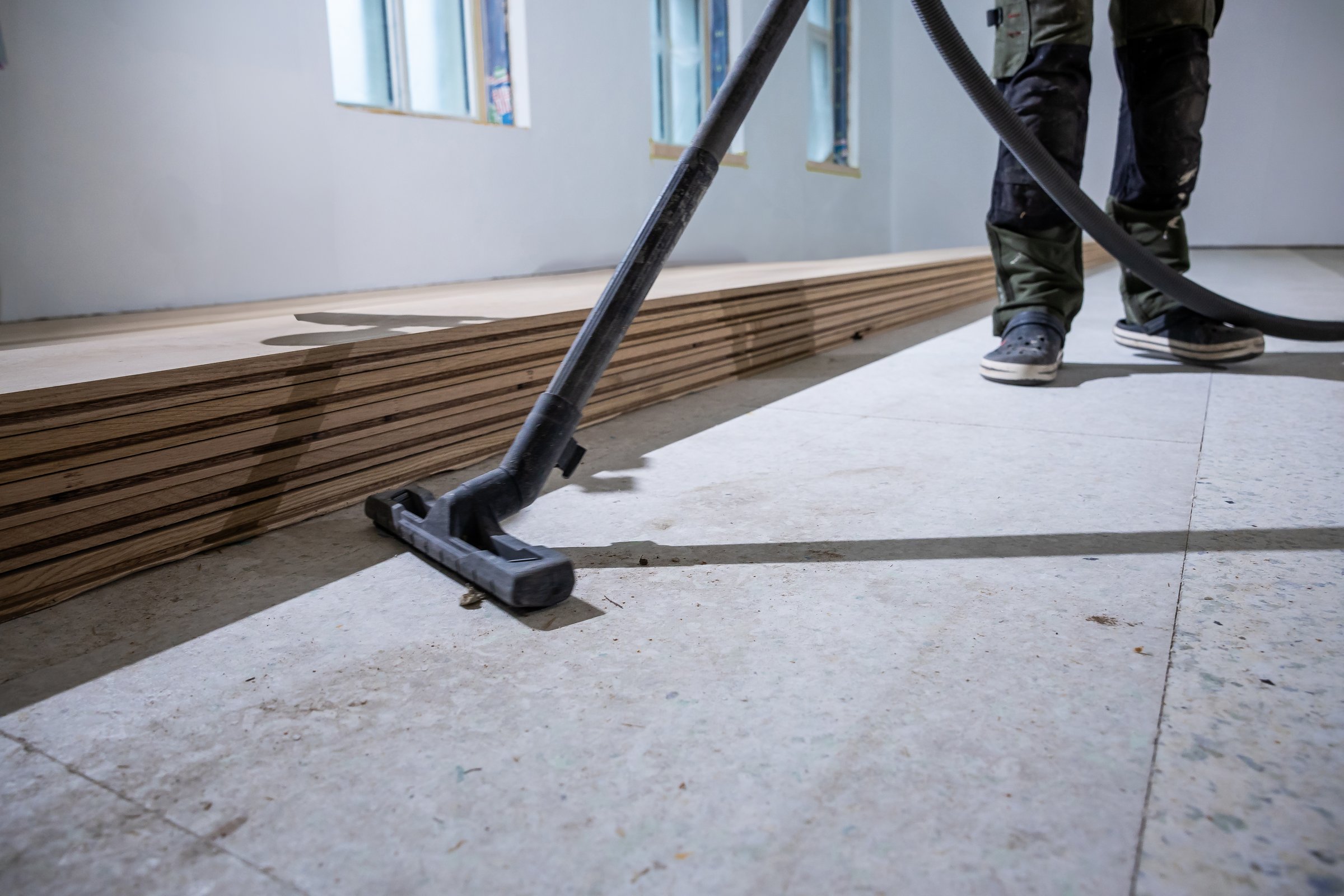 Un operaio pulisce il sottopavimento con un aspirapolvere in una stanza ben illuminata. Assi di legno ordinatamente impilate sono visibili, con luce naturale che filtra dalle finestre.