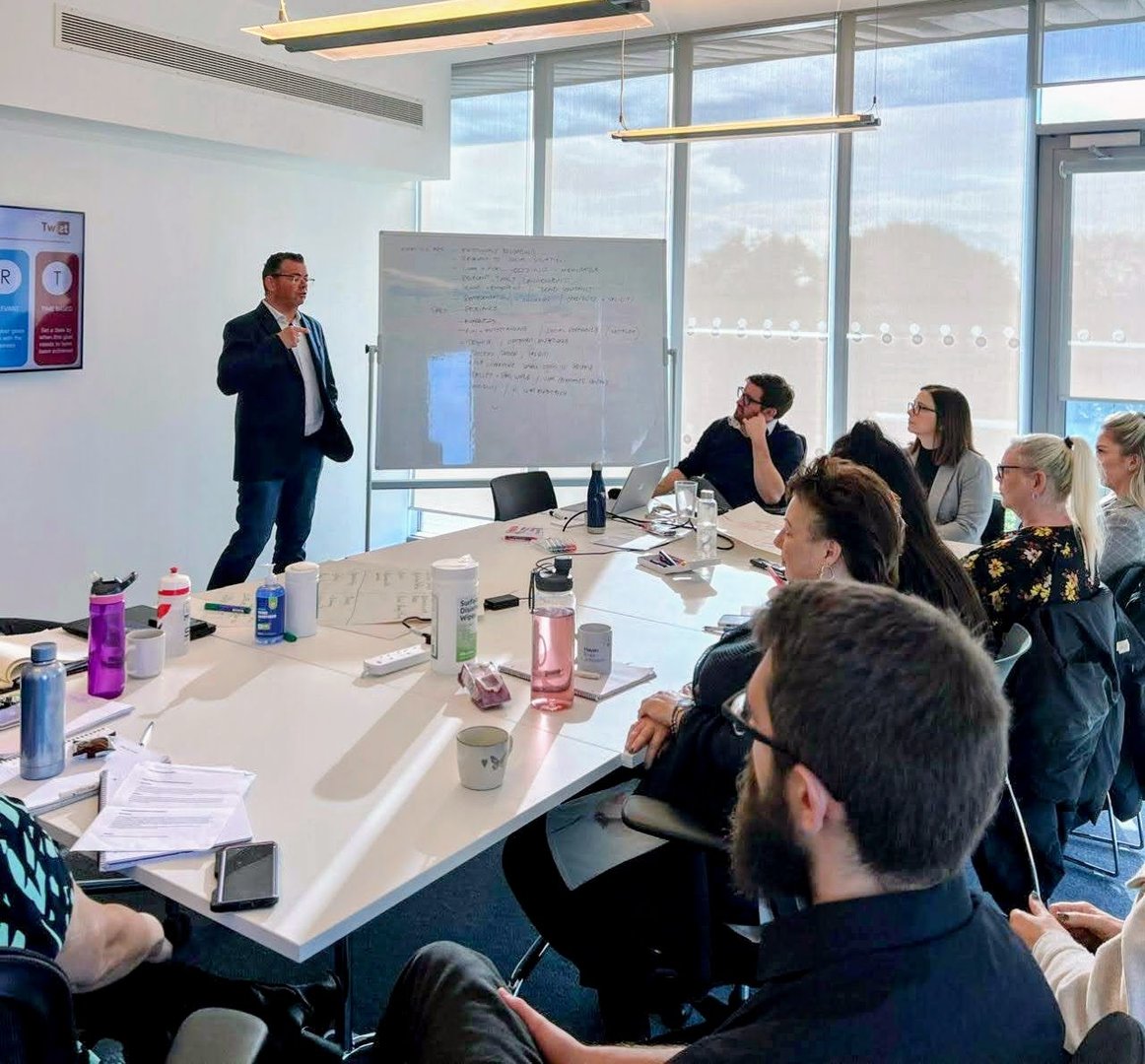 A man presents in a conference room to a group seated around a table with papers and water bottles. A whiteboard is visible.