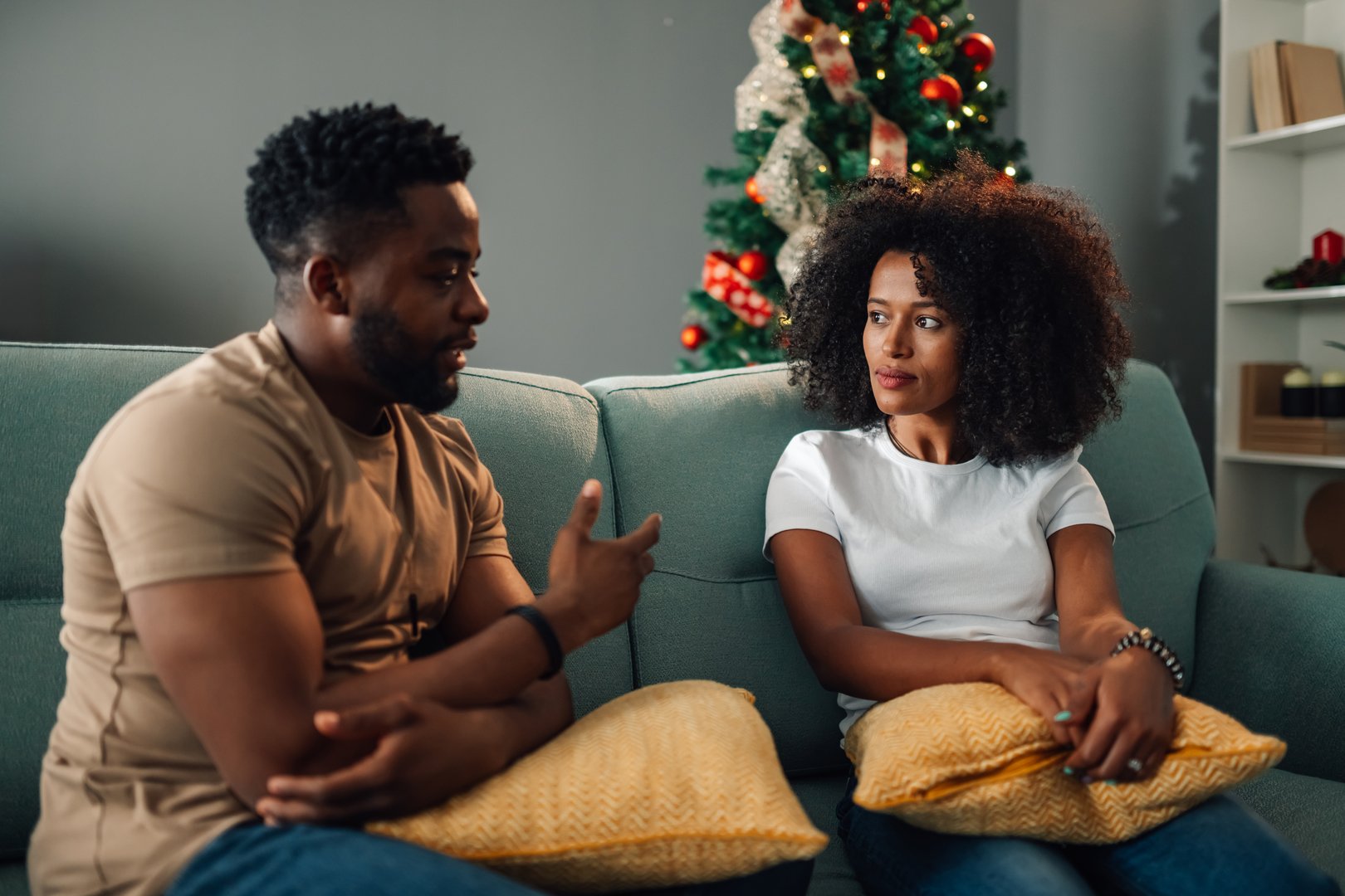 African american couple sits on a sofa in animated conversation, framed by a beautifully decorated Christmas tree, illustrating lively exchange and warmth during a festive, cozy home setting.