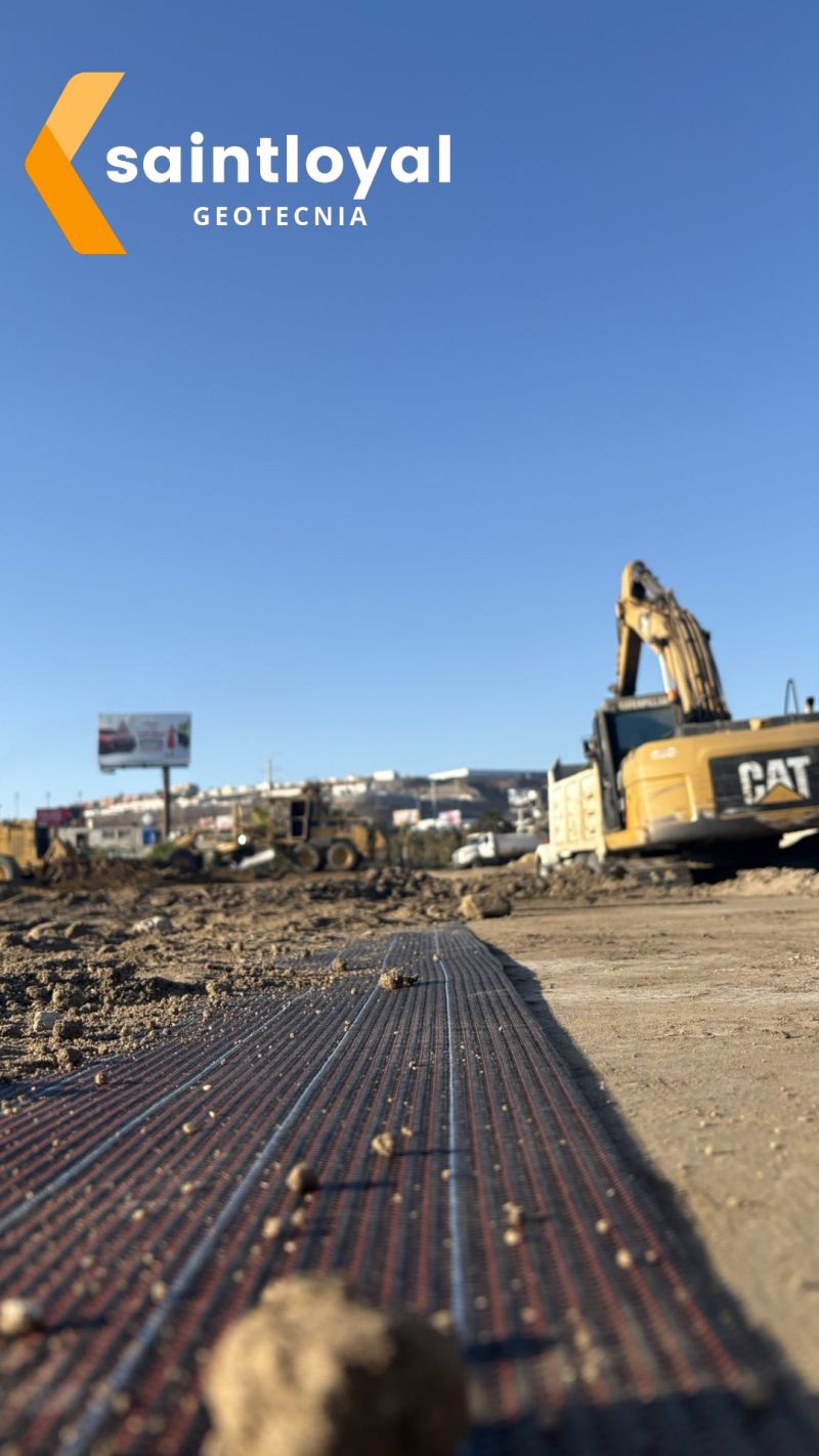 Construction site with heavy machinery and geotextile sheet in foreground, under a clear blue sky.