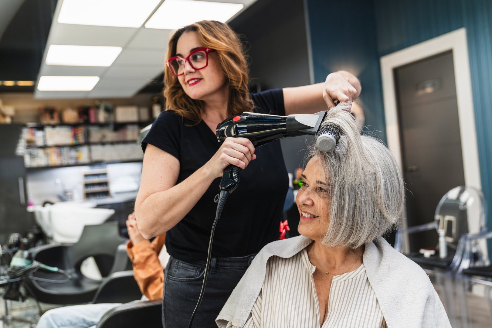 Professional hairdresser drying and styling a smiling senior woman's gray hair with a brush and hairdryer at a beauty salon