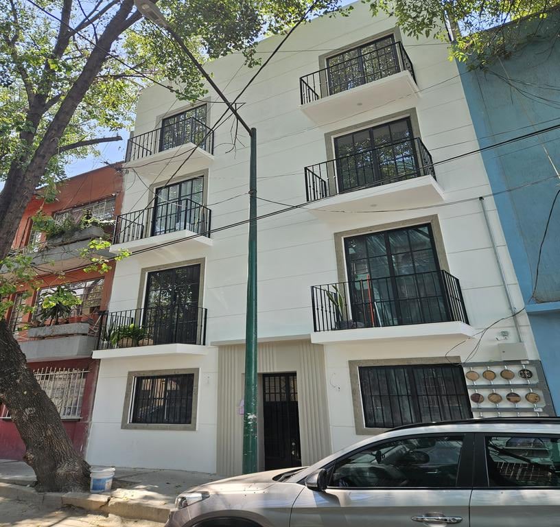 Four-story white building with balconies, trees lining the street, and a parked car in front.