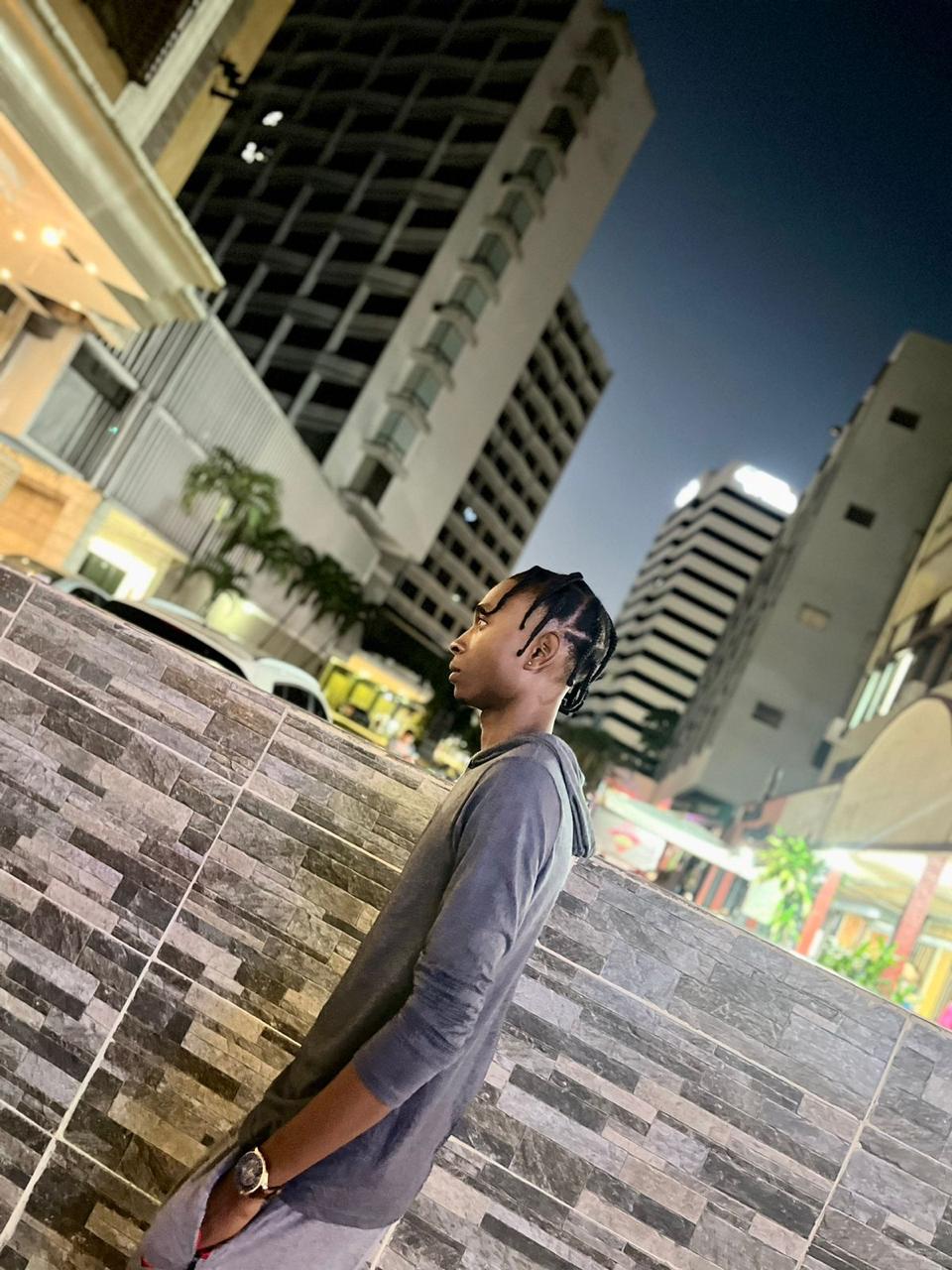 A person in a hoodie stands outdoors near a stone wall with tall buildings in the background at dusk.