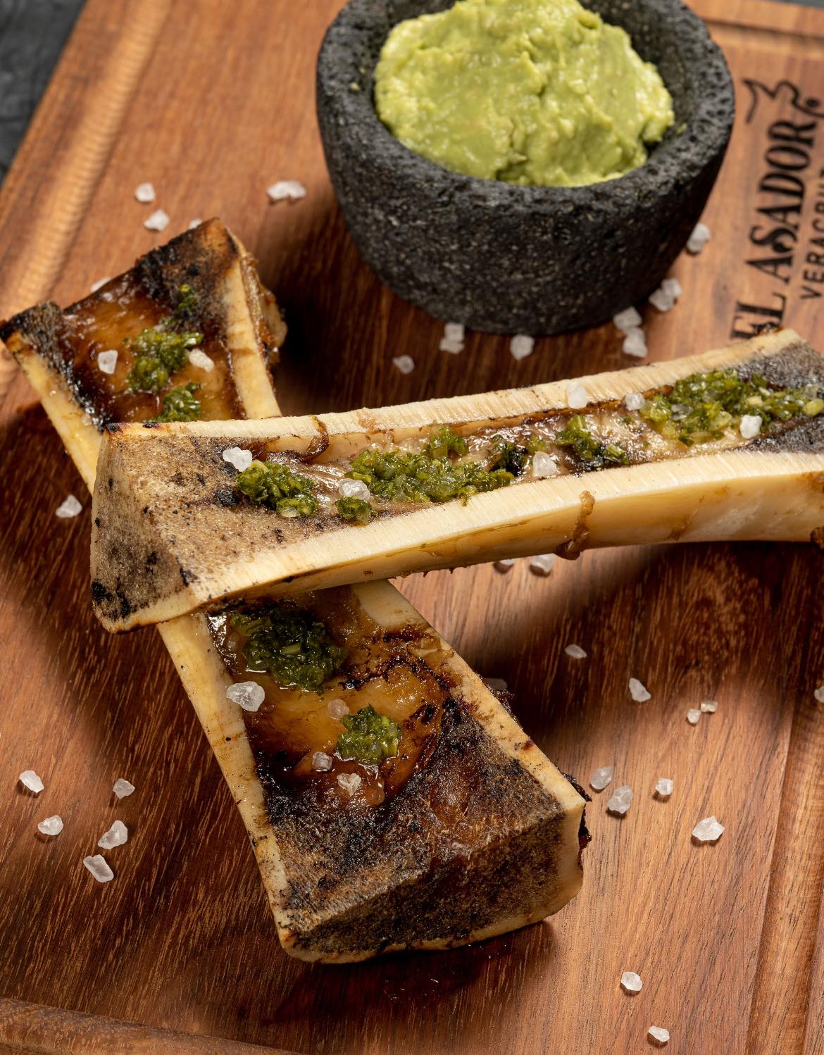 Grilled bone marrow with seasoning on a wooden board, served with a mortar of guacamole on the side.