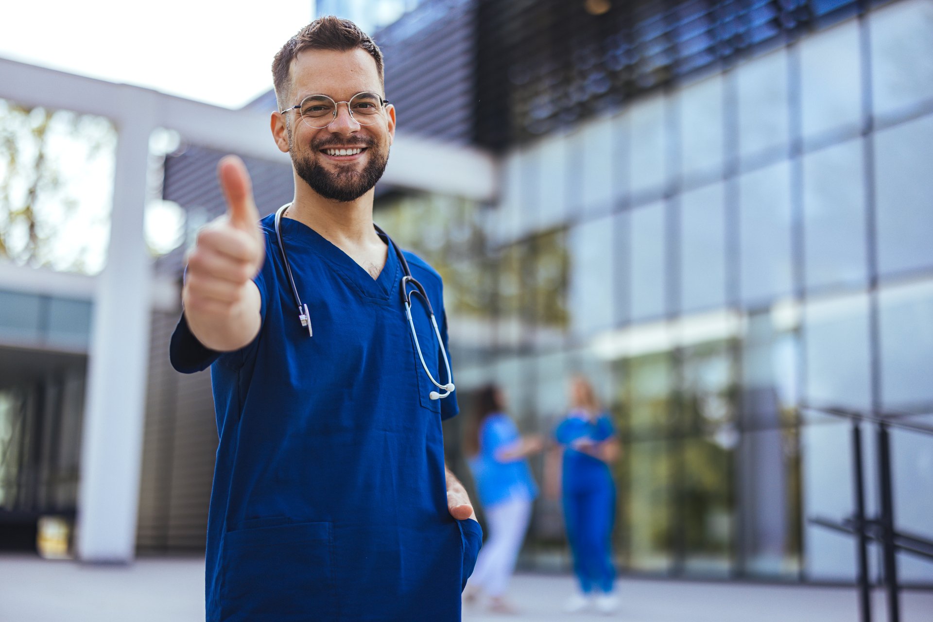 Handsome man with beard wearing blue male nurse uniform doing happy thumbs up gesture with hand. Approving expression looking at the camera showing success.