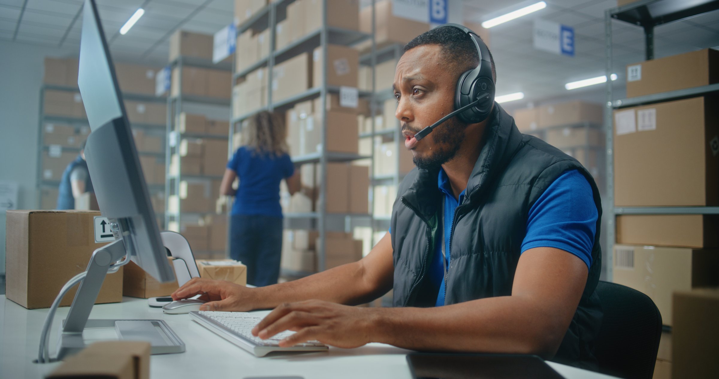African American logistics coordinator wearing headset chats with customer on call, uses computer, works in post office or E-commerce store storage. Sorting center workers carrying cardboard boxes.