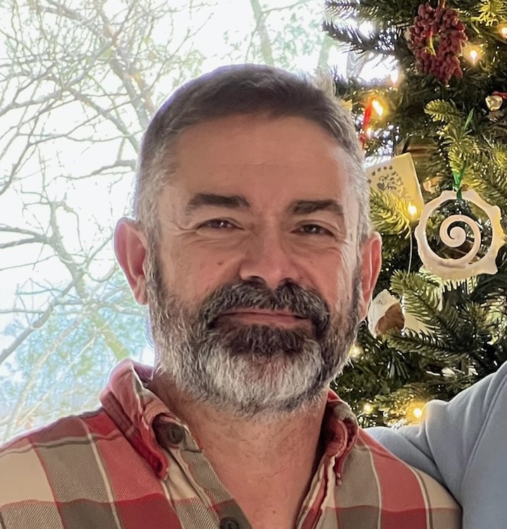 A man with a beard wearing a plaid shirt stands next to a decorated Christmas tree with lights and ornaments.