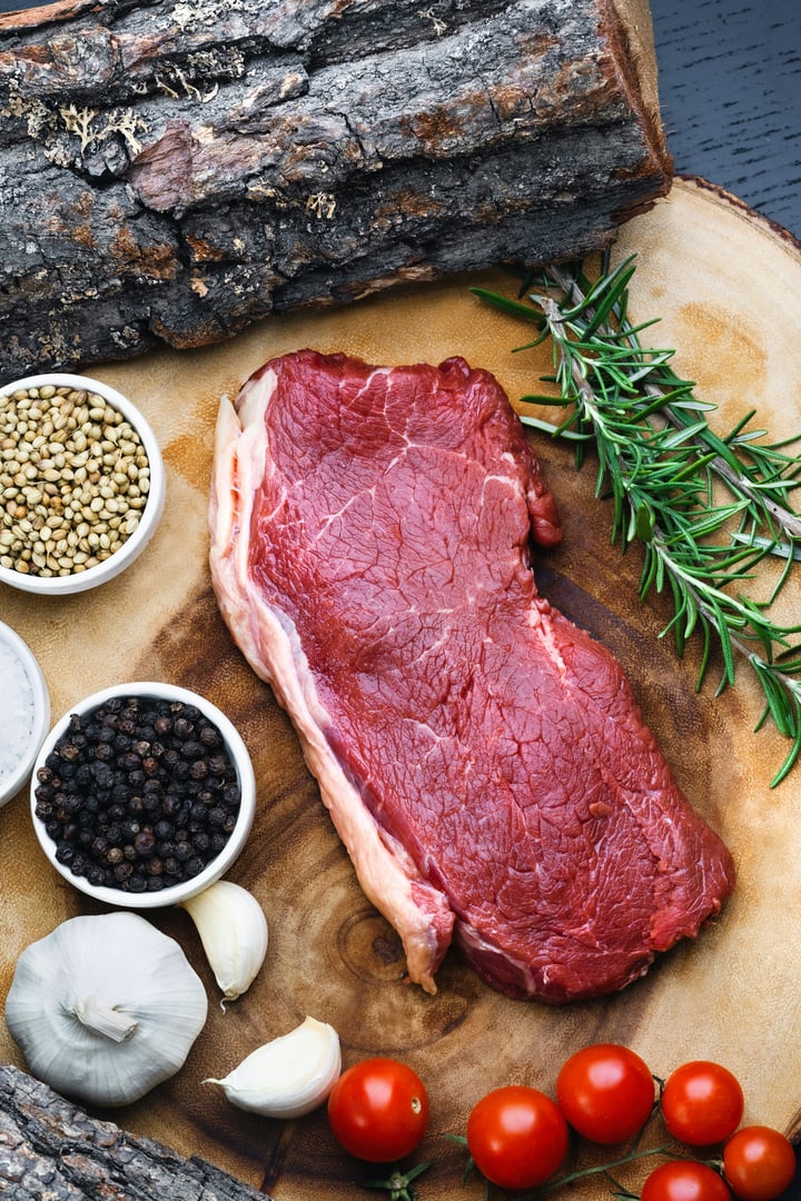 Raw steak with rosemary, cherry tomatoes, garlic, peppercorns, and coriander seeds on a wooden surface.
