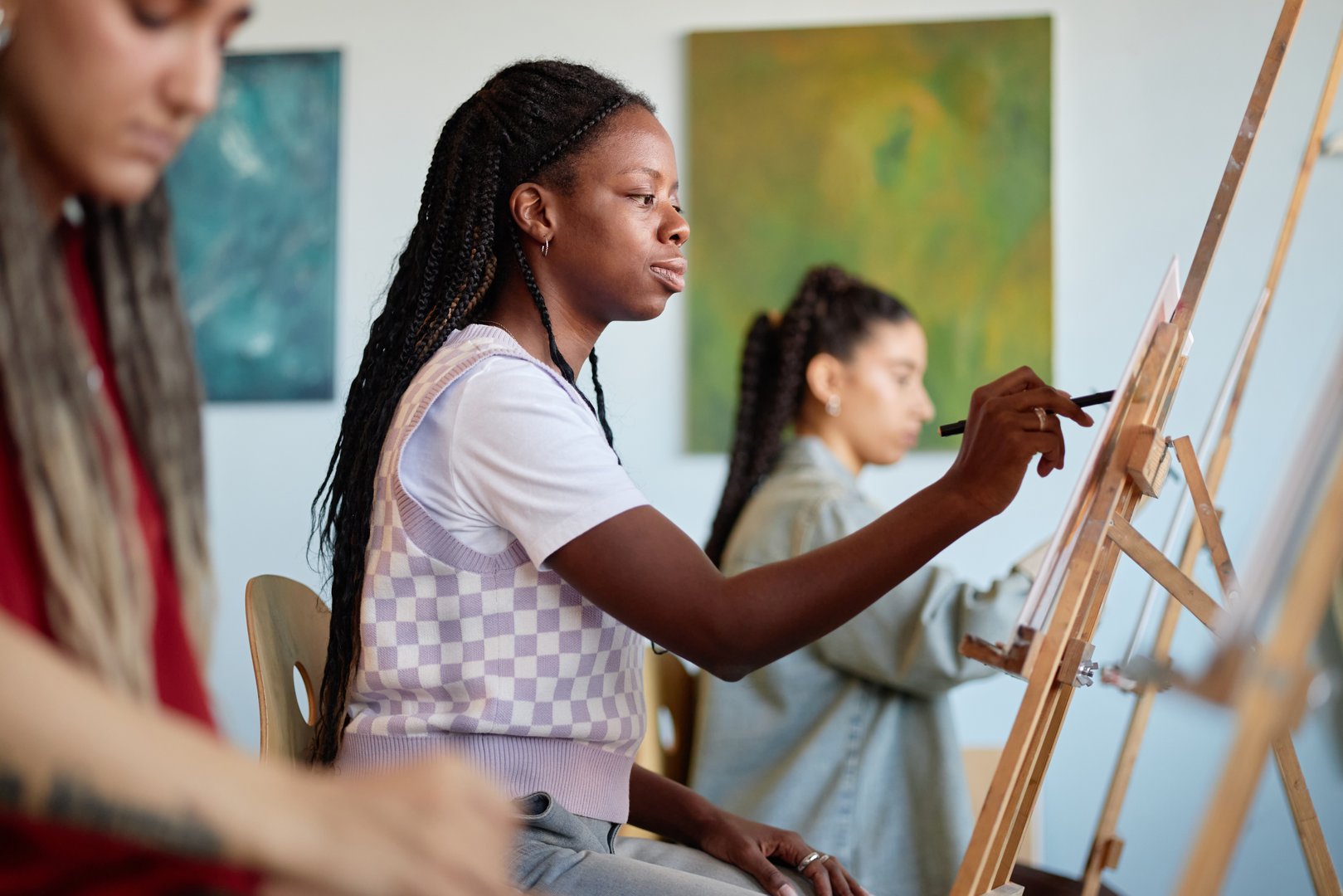 Black young adult woman sitting at easel painting on canvas in art studio, surrounded by other young adults engaged in creative activity, focused on artistic process