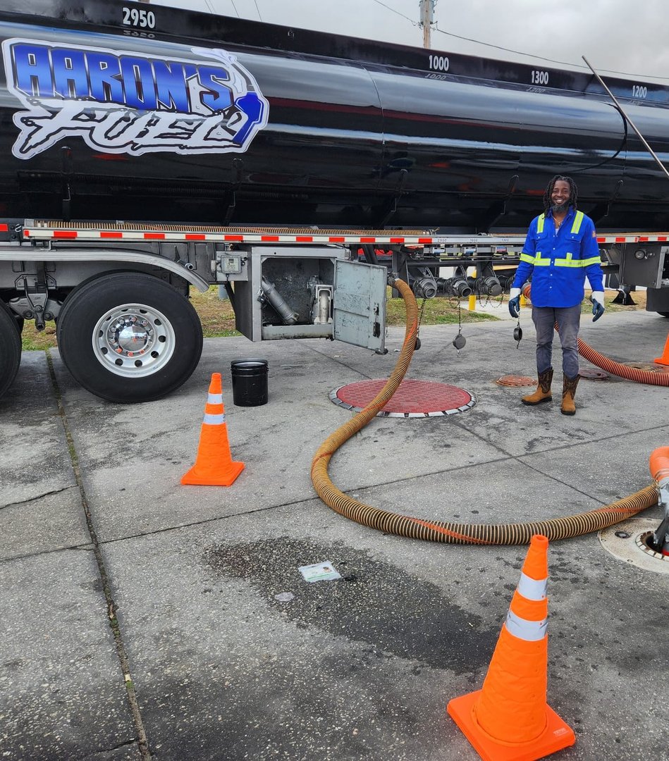 Worker in safety gear refueling at a gas station with a tanker truck and orange cones surrounding the area.