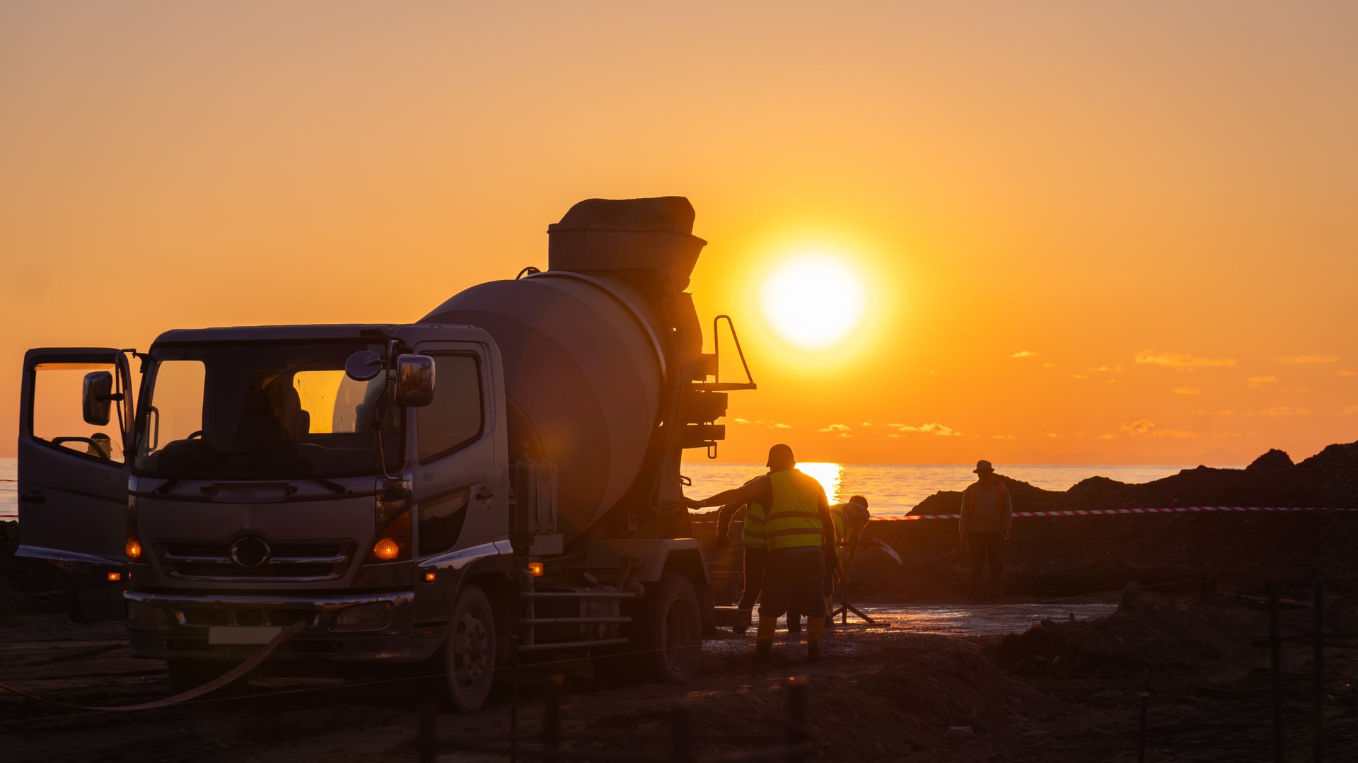 Close-up at sunset on the seashore of a concrete mixer truck with people in work uniforms
