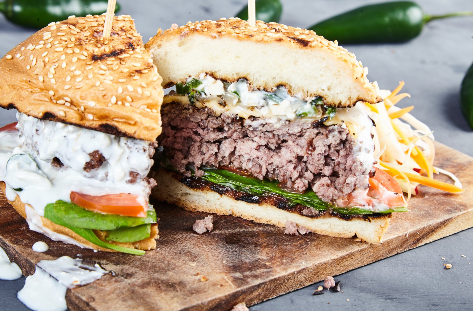 Gourmet homemade burger with beef, fried spicy jalapenos, fresh spinach, sliced tomatoes, cheese, grilled bun and sauce of yogurt and blue cheese. With coleslaw on wooden board. Jalapenos behind