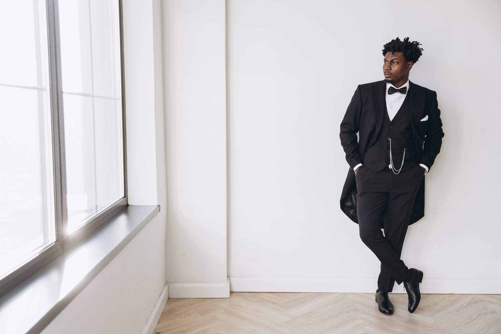 Stylish groom wearing black suit, bow tie and pocket watch chain posing in a white room with parquet floor and large window