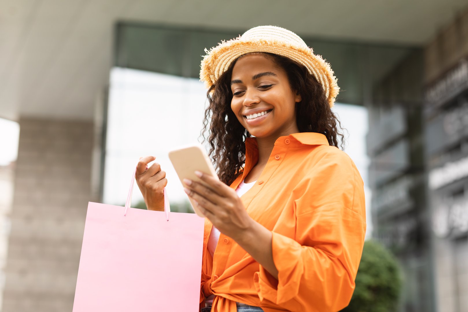 Happy young pretty african american woman in casual, hat with many bags with purchases typing on smartphone, use banking on city street. New online shopping app, shopaholic enjoy sale and fashion blog