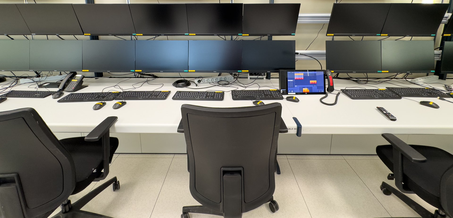 An empty control room with multiple computer workstations. There are six monitors at each station. The monitors are all black and are connected to a white desk.