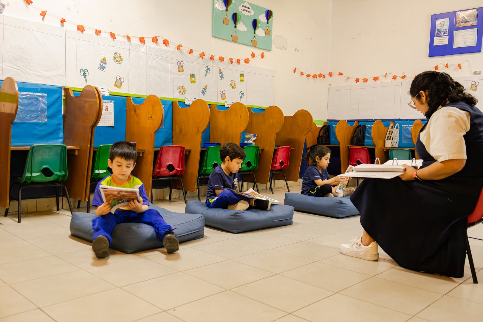Children reading on cushions in a classroom with a teacher sitting nearby, surrounded by colorful decor and dividers.