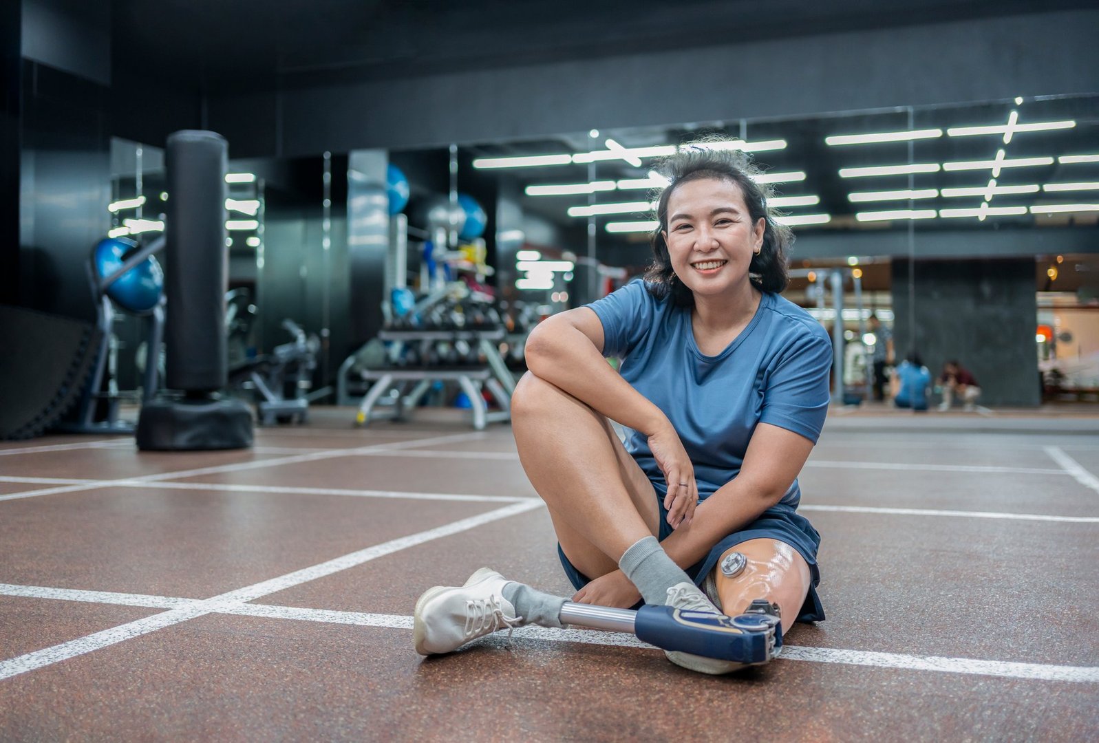 portrait cheerful asian female with prosthetic leg sitting on floor and posing to camera while resting after workout at indoors gym