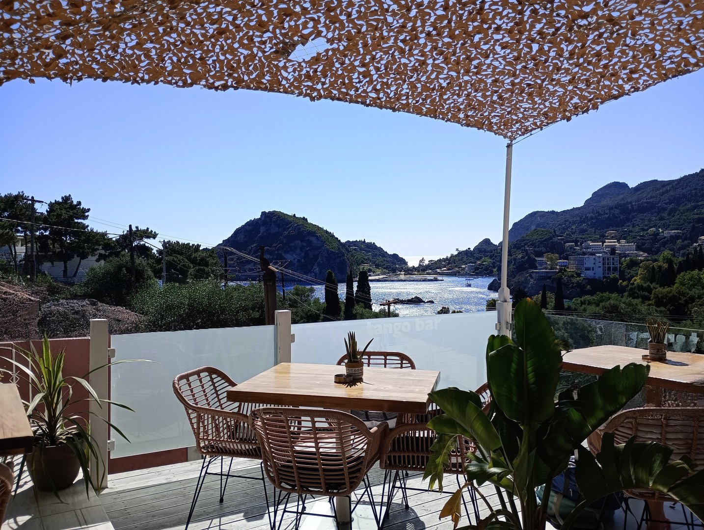 Taken in Paleokastritsa, Corfu, Greece, this image captures a stylish outdoor cafe terrace with wooden tables, wicker chairs, potted plants, and a shaded canopy. Beyond the glass barrier, the Ionian Sea sparkles under a clear blue sky, framed by hills and coastal buildings. No people are visible, making this perfect for Mediterranean design content, lifestyle themes, travel visuals, or hospitality industry use. Natural light and layered depth make it suitable for editorial and promotional projects.