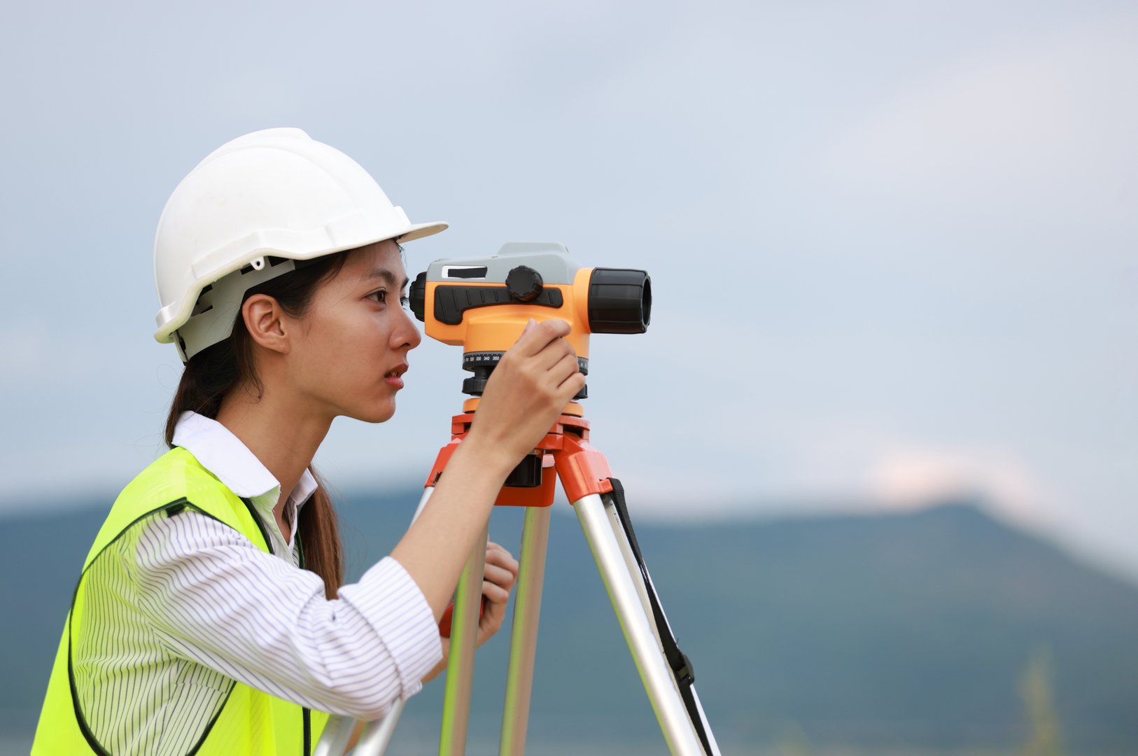 Portrait beautiful Asian construction engineer with a green safety vest hard hat and level survey camera.Engineer peering through a theodolite