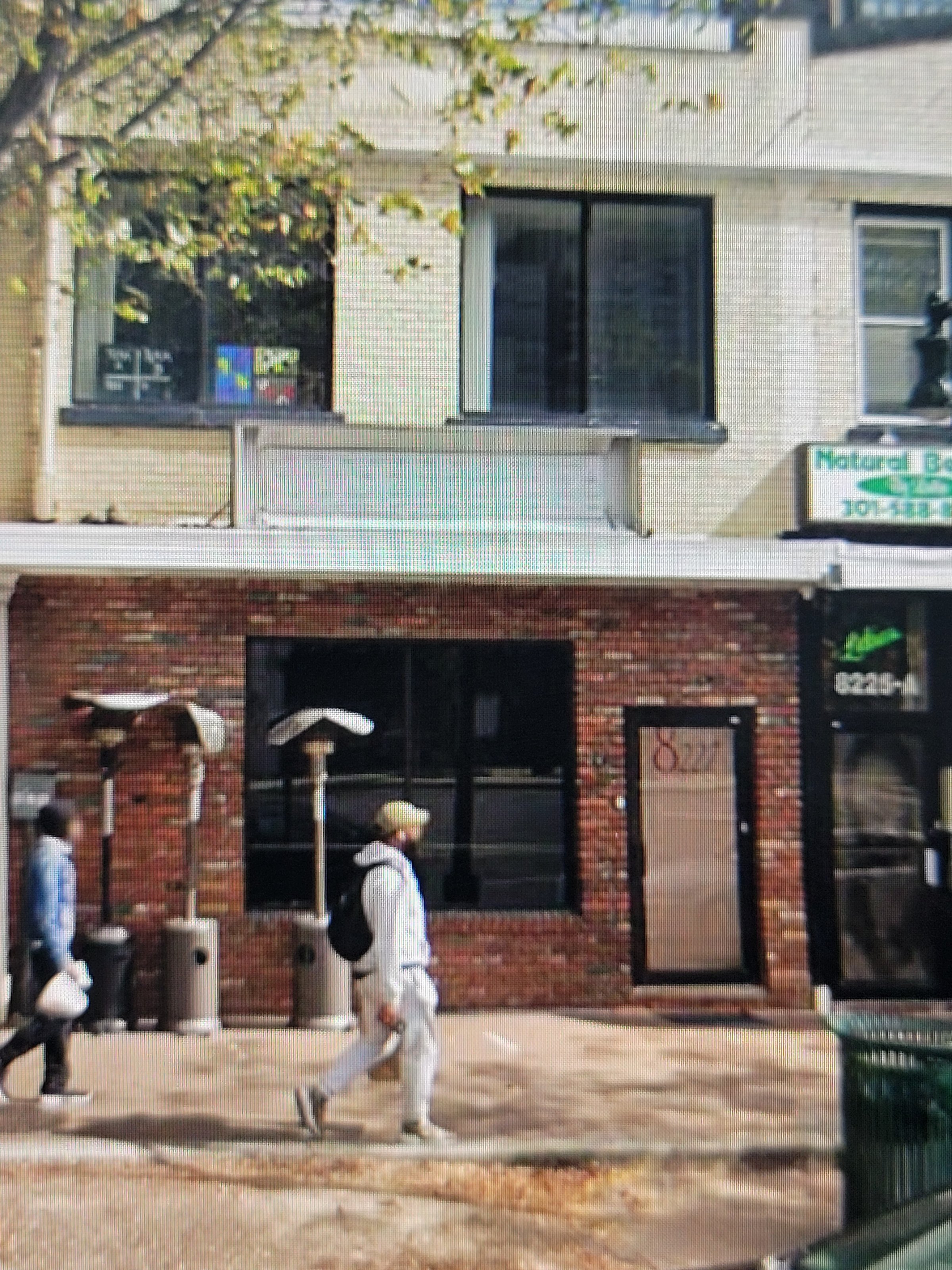 Street view of a brick building with large windows, a tree above, and a person walking on the sidewalk carrying a bag.