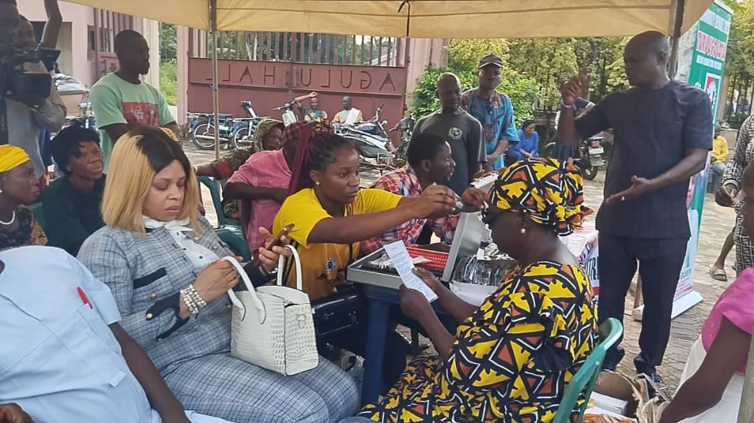 A group of people, including a woman applying makeup, gathered under a tent with a Nigerian cultural setting.