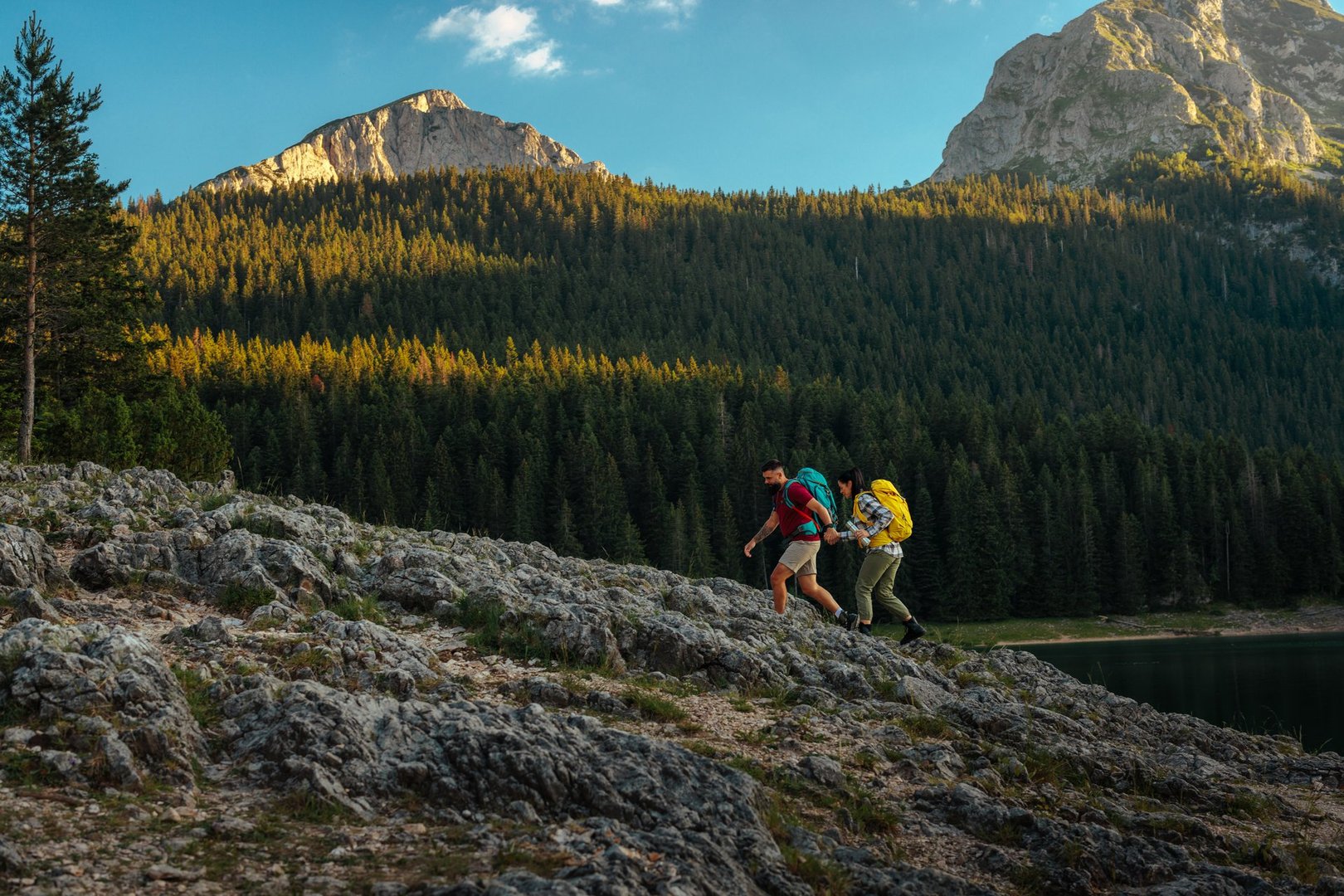 Wide shot of a couple hiking through rocky terrain holding hands as they walk uphill with a forest and mountains in the background.