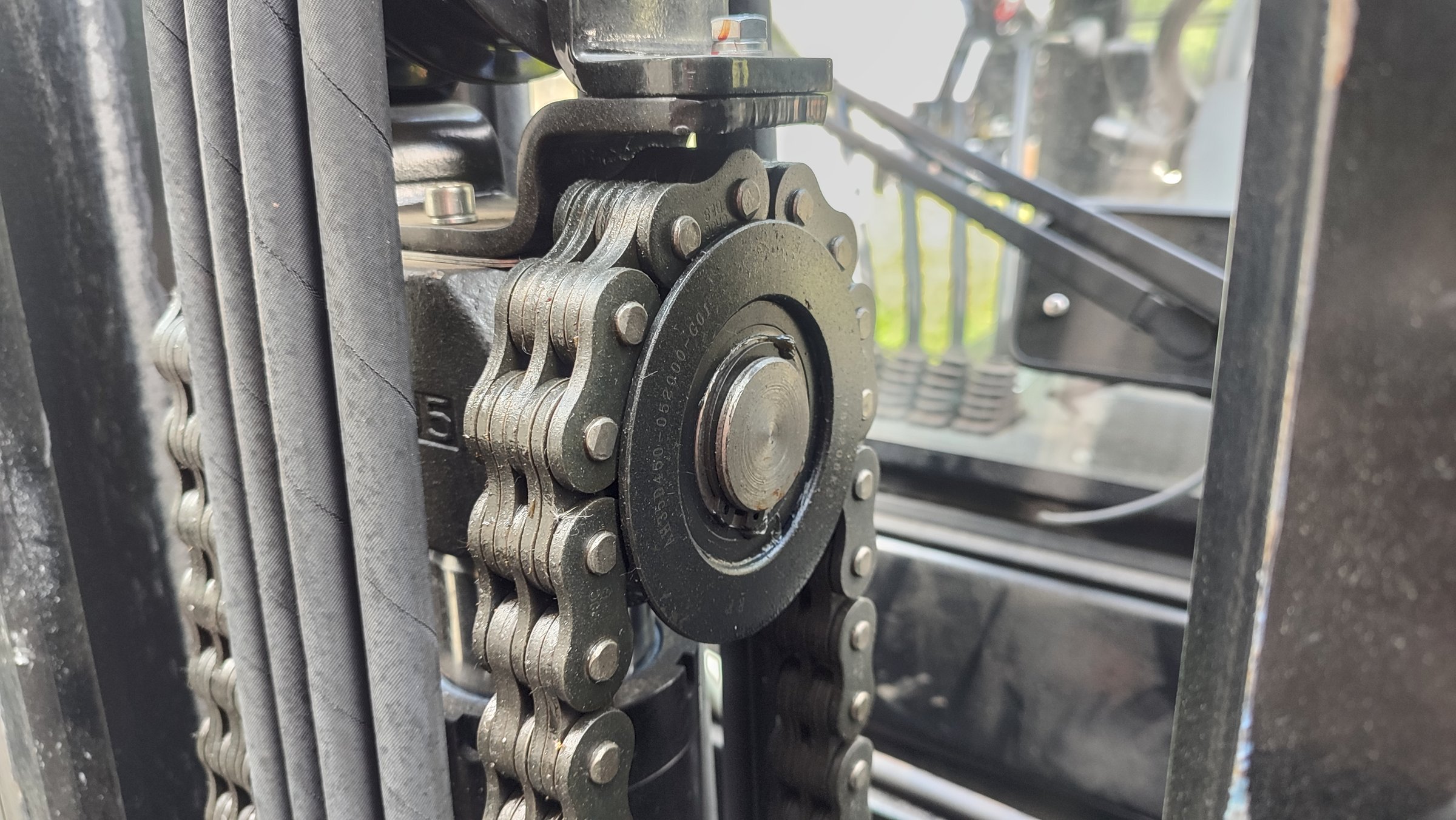 Close-up of a forklift mast showing the chain mechanism, pulley, and hydraulic hoses in an industrial setting.